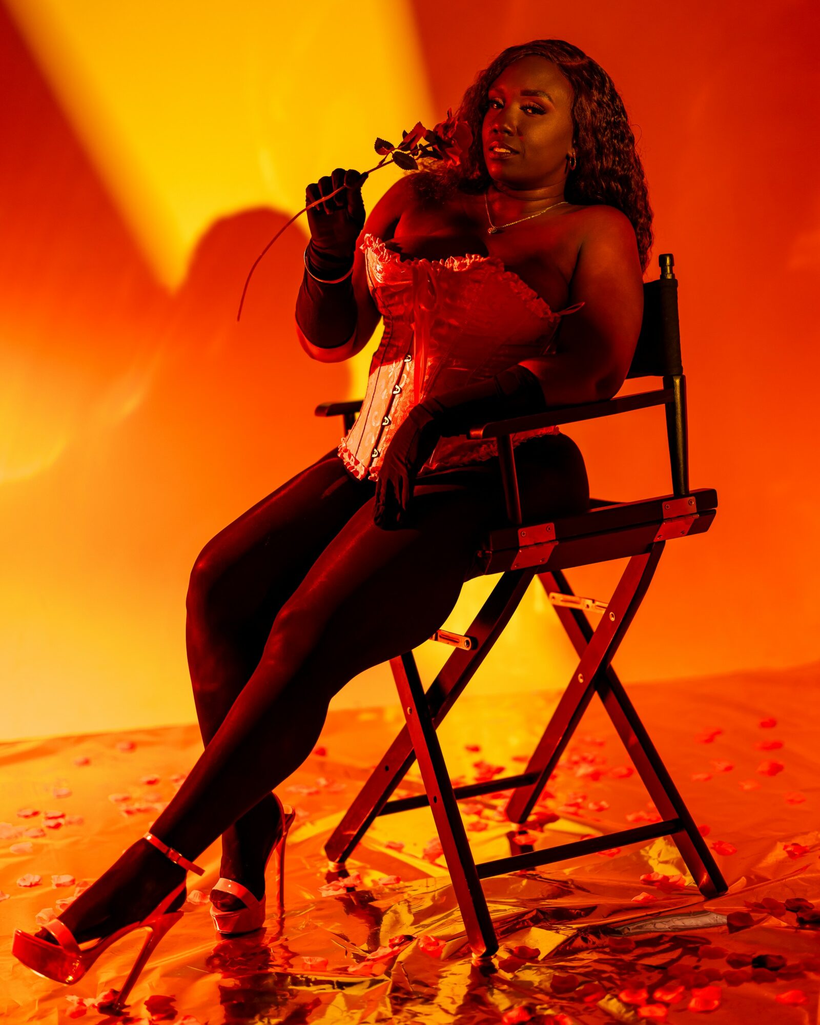 Woman sitting on a chair holding a flower, with warm orange and yellow background, wearing a red outfit and high heels.