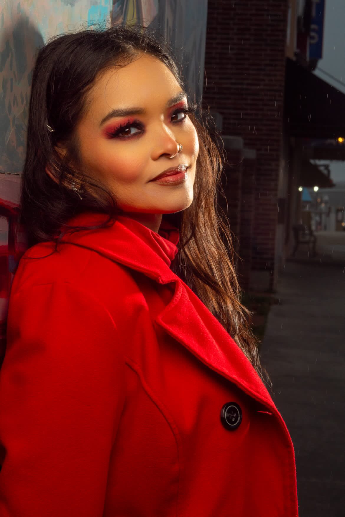 Young woman with long hair wearing a red coat, smiling, in an urban setting at night.
