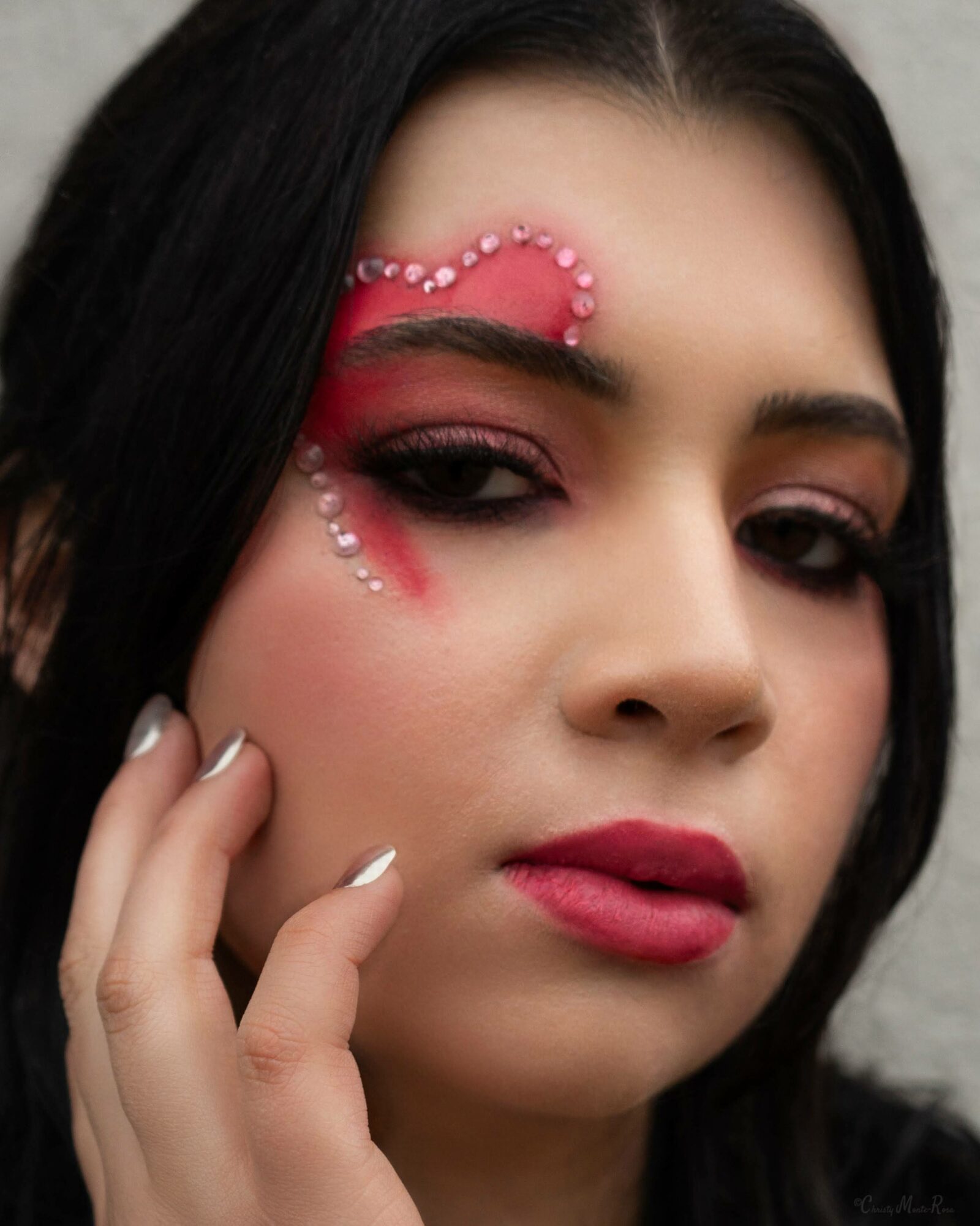Close-up of a woman with makeup and decorative rhinestones around her eye, touching her face gently.