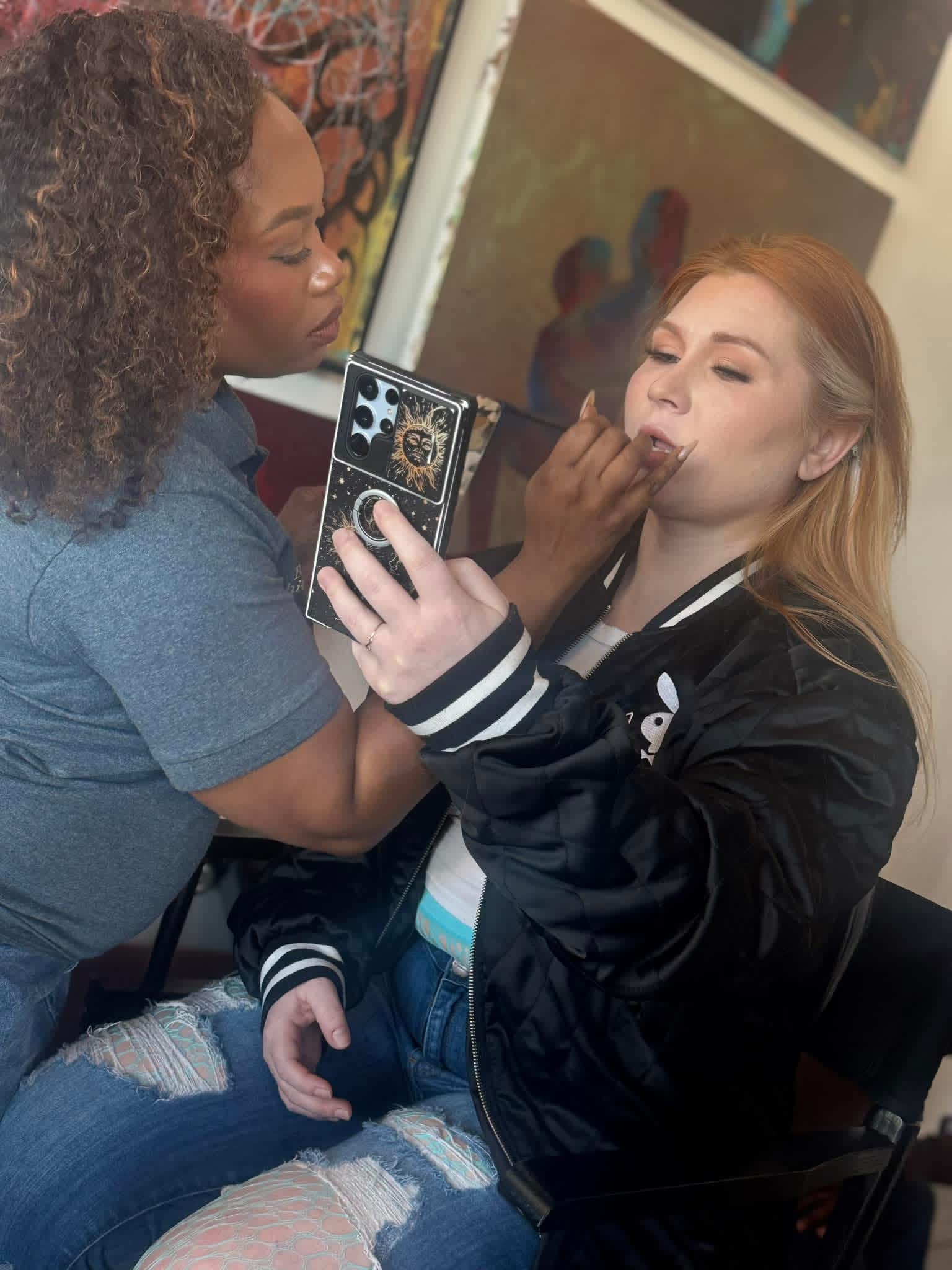 Two women, one applying makeup to the other, in an indoor setting with artwork in the background.