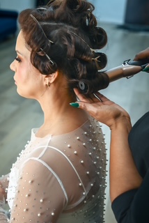 Woman with styled hair and pearl-embellished sheer top, getting her hair curled by a stylist, side view.