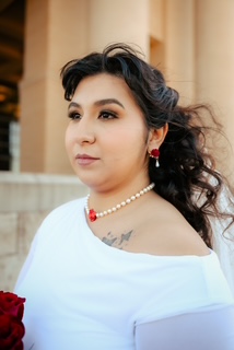 Woman with dark curly hair wearing pearl and red jewelry, looking to the side, in front of a beige building.