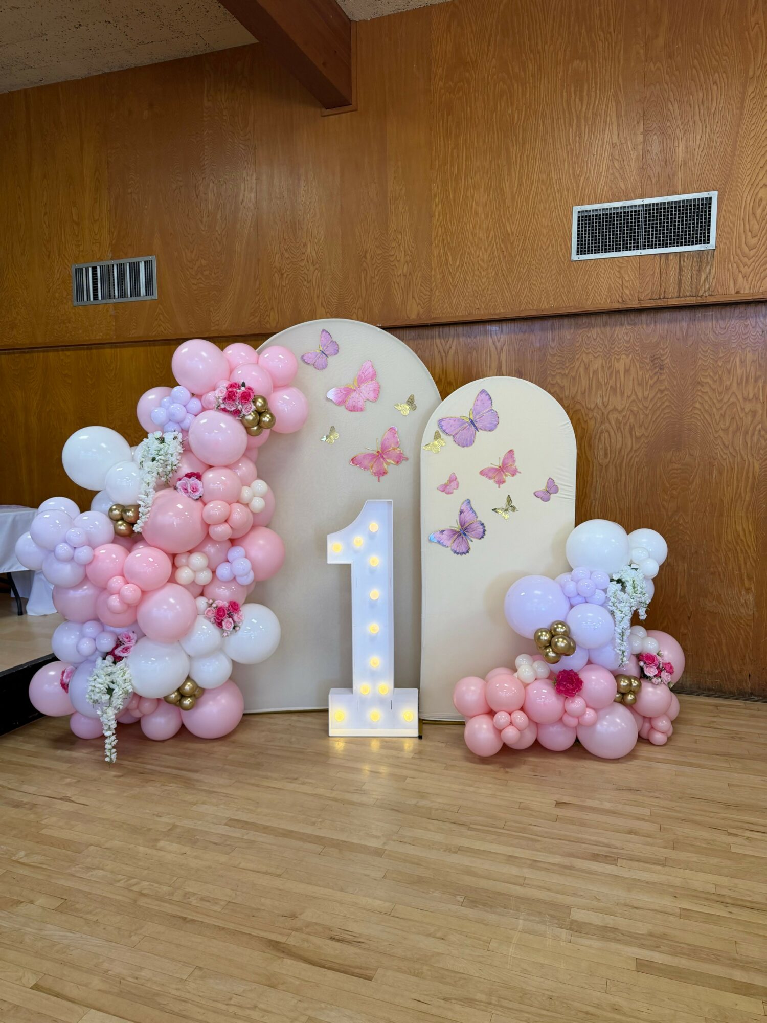 Decorative arrangement with pink, white, and purple balloons, and a large letter 'I' with lights, on a wooden floor.