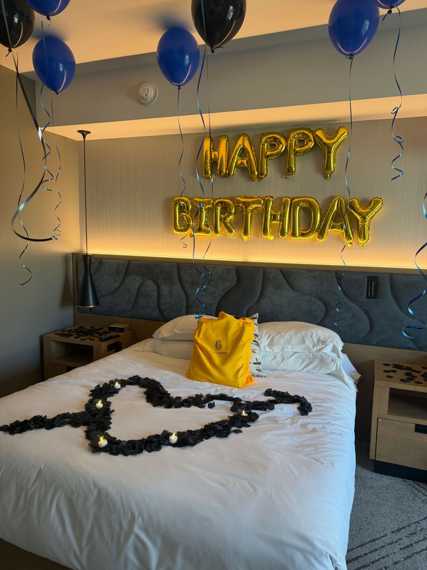 Decorated bedroom with birthday balloons, a necklace on the bed, and a yellow cloth. Wall has gold 'Happy Birthday' balloons.