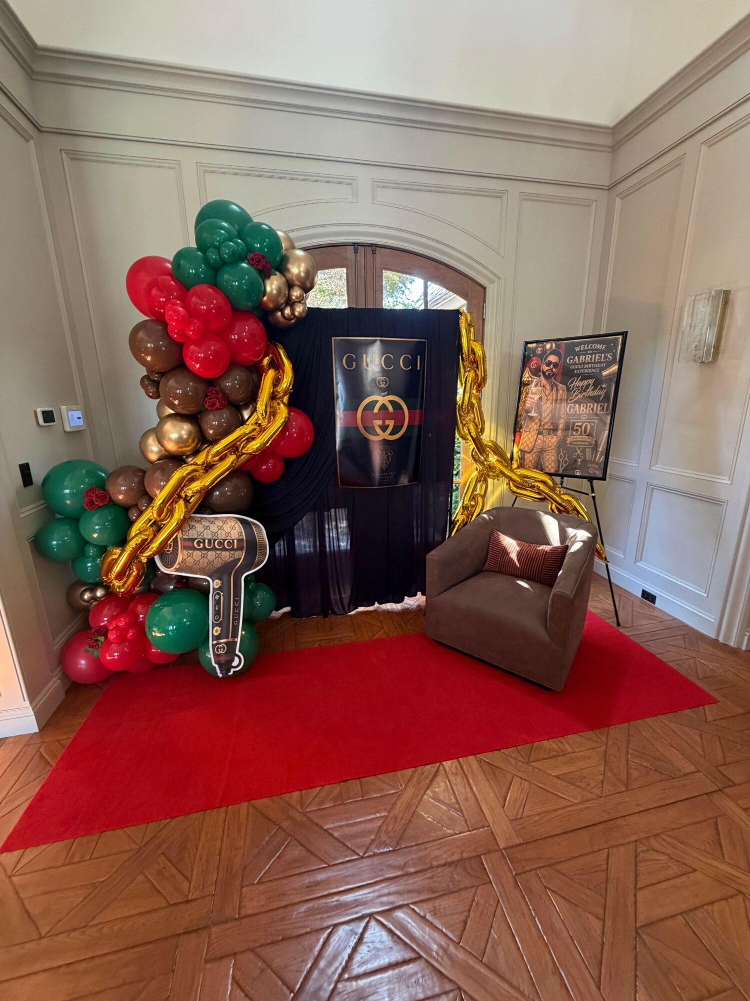 Decorative display with balloons, a black backdrop, and a gold balloon sculpture in a room with wooden flooring and a red carpet.