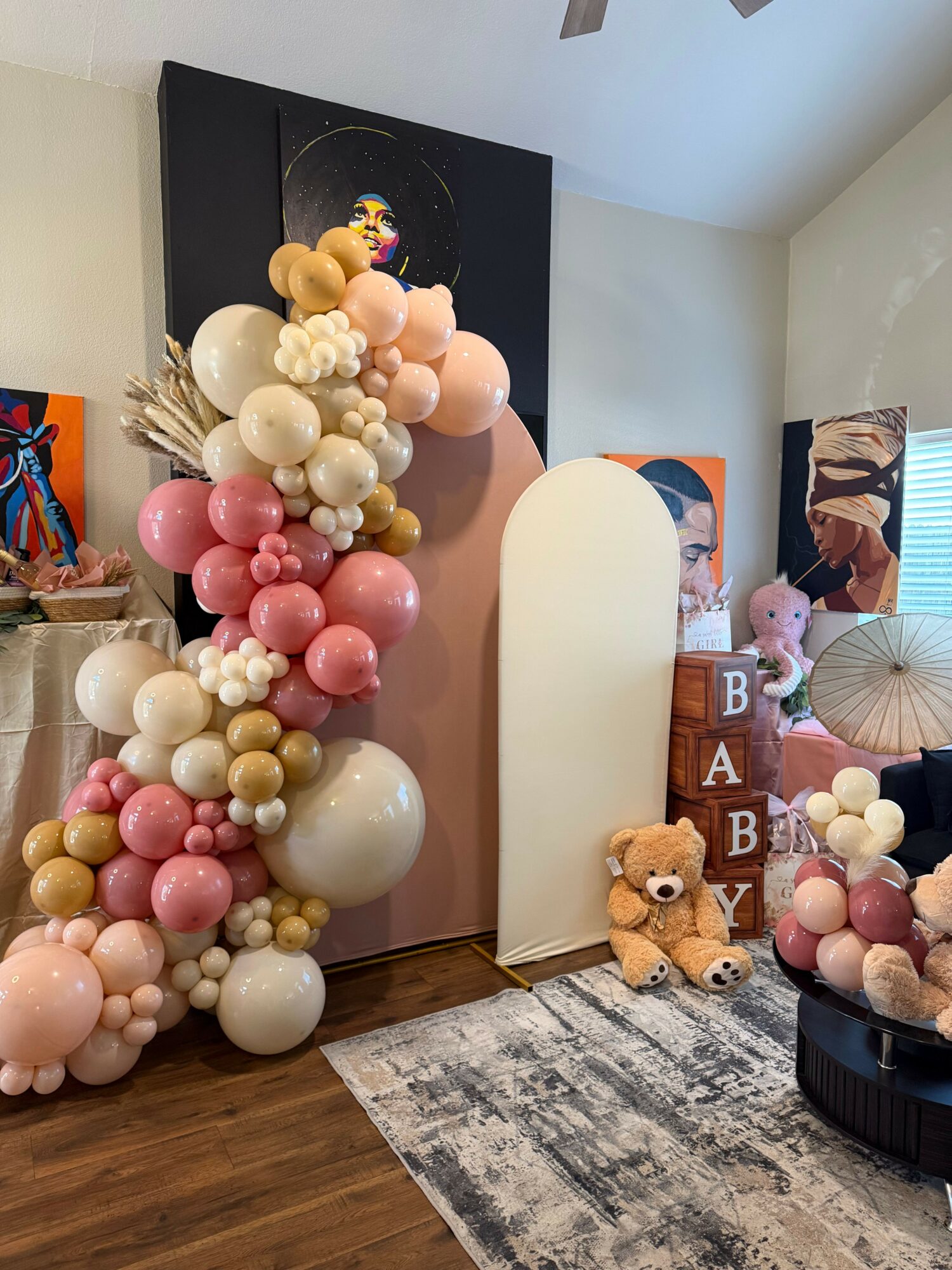 Decorative balloon arrangement with pink, white, and gold balloons, teddy bears, and a tray of sweets on a wooden floor.