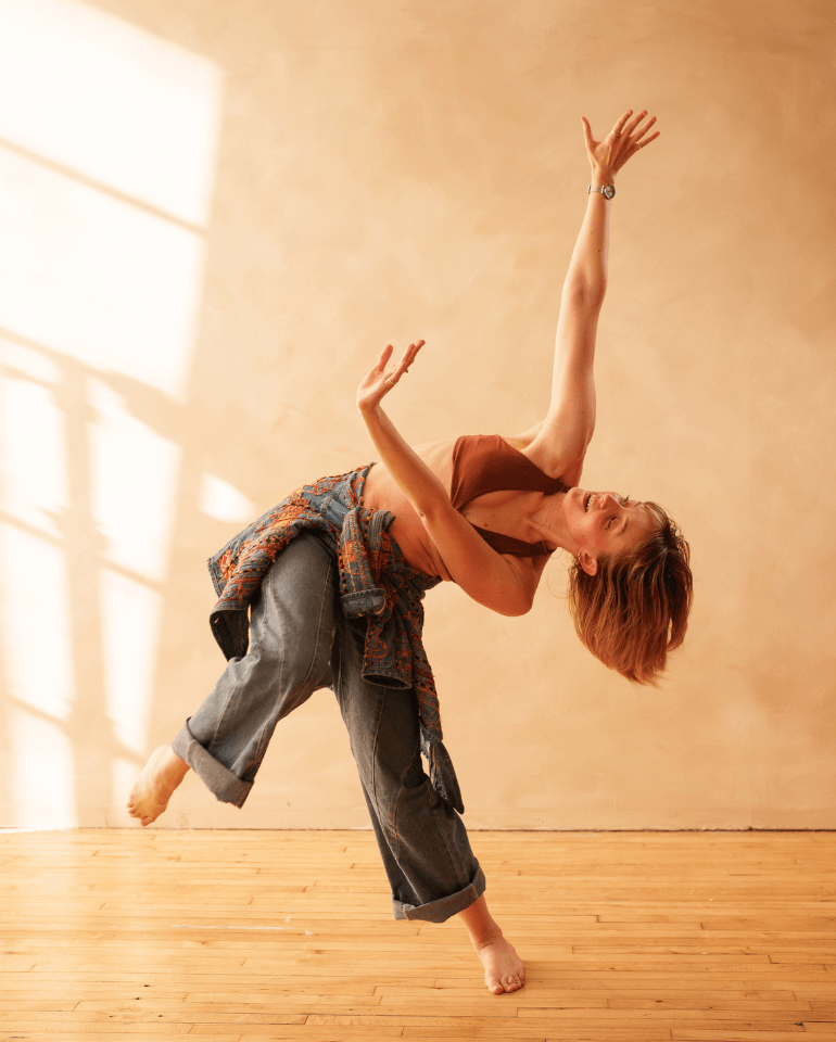 A woman performs a dance move with one arm raised and the other bent, standing on one leg in a studio.