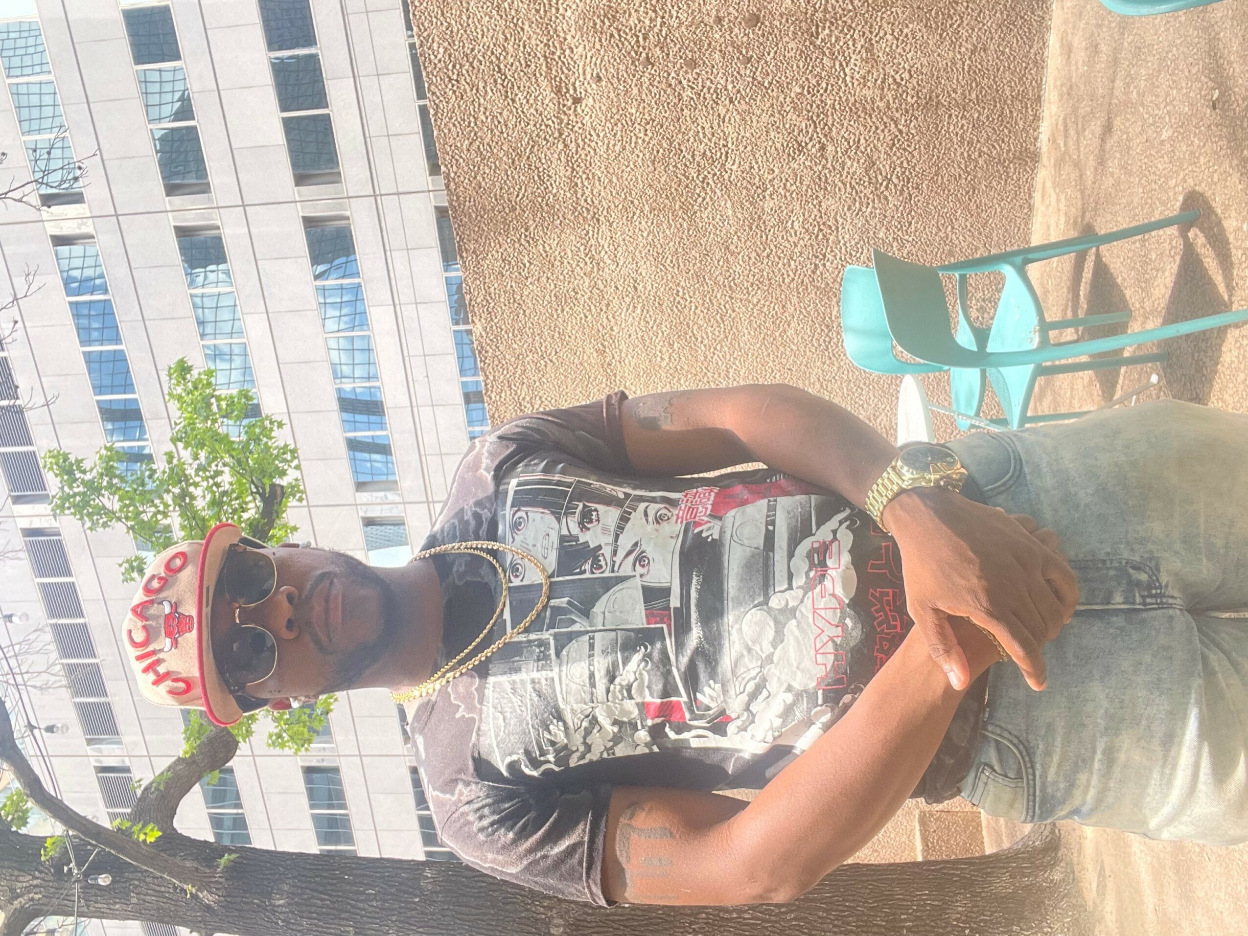 Person wearing a cap, sunglasses, and casual clothing standing outdoors with buildings and chairs in background.