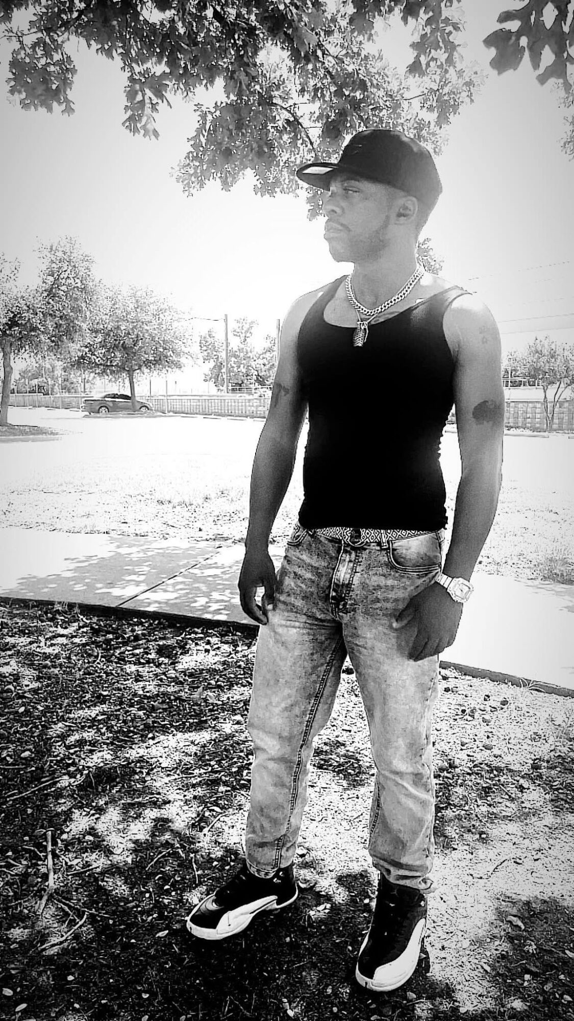 Young man standing outdoors wearing a cap, sleeveless shirt, jeans, and sneakers, with trees and sunlight in background.