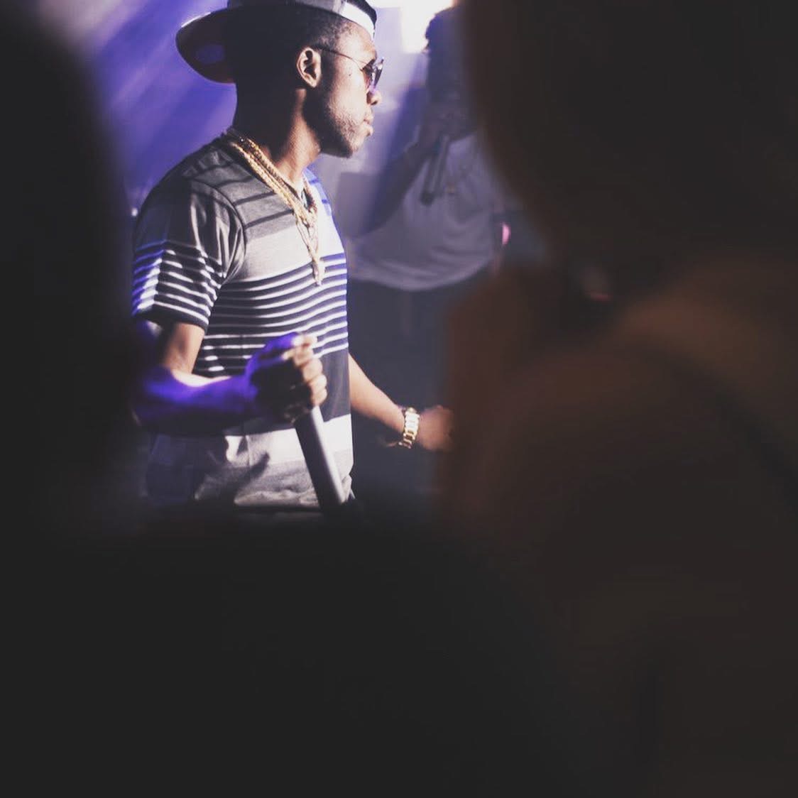 Man wearing a striped shirt, hat, and glasses holding a microphone in a dimly lit setting.