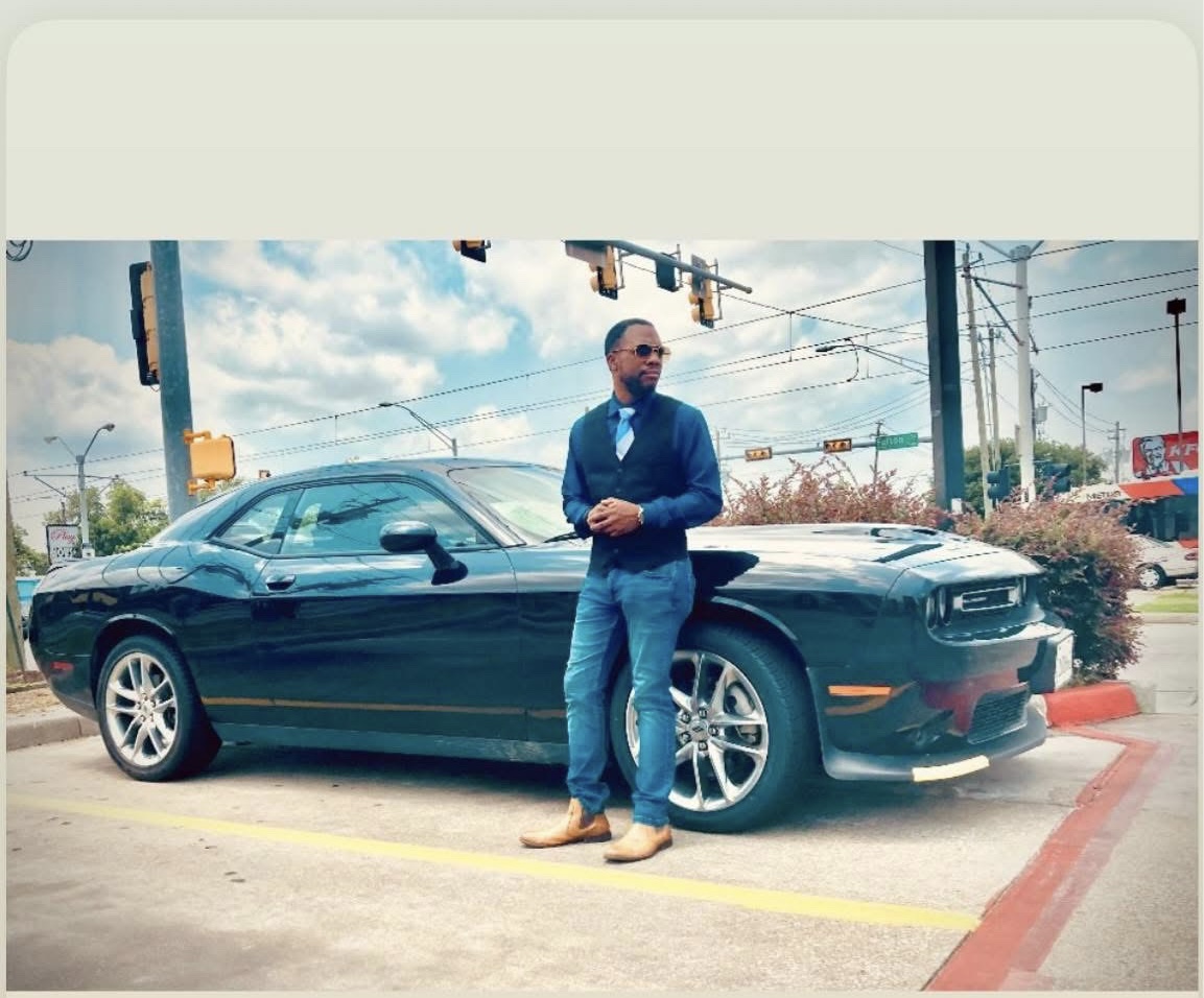 Man in blue blazer and jeans standing next to a black sports car at an intersection with traffic lights and power lines