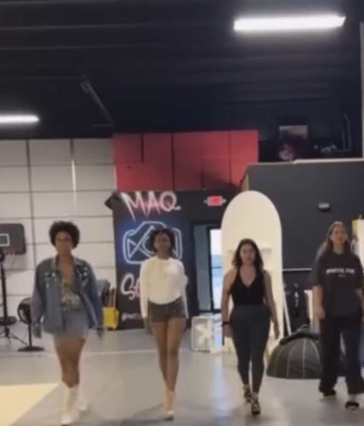 Four people walking in an indoor space with a black ceiling and illuminated signs, some wearing casual clothing.