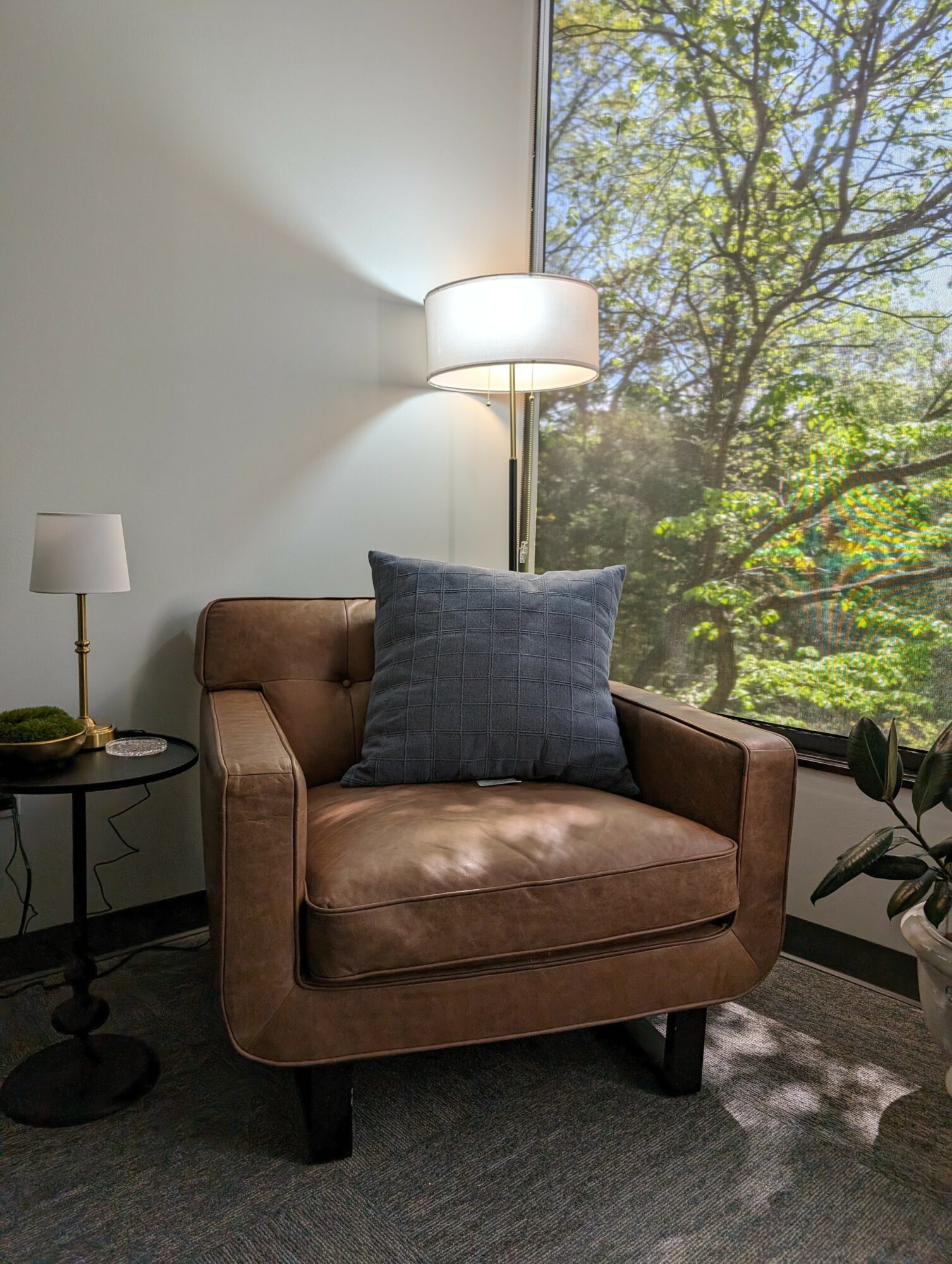 Brown armchair with a blue pillow, next to a small side table with a lamp, in front of a window with trees outside.