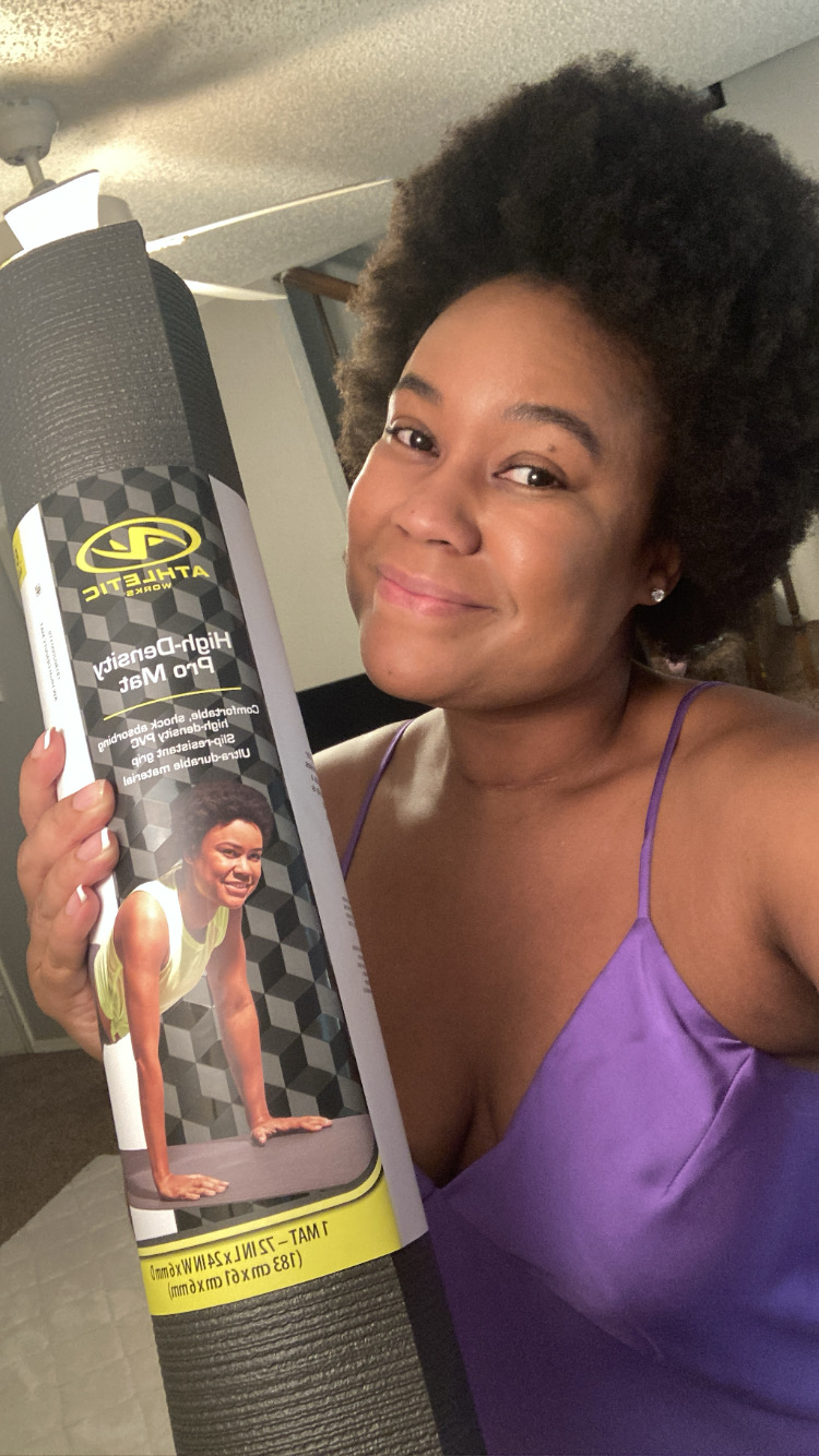 Woman with natural hair smiling, holding a yoga mat with a label, wearing a purple top, indoors.