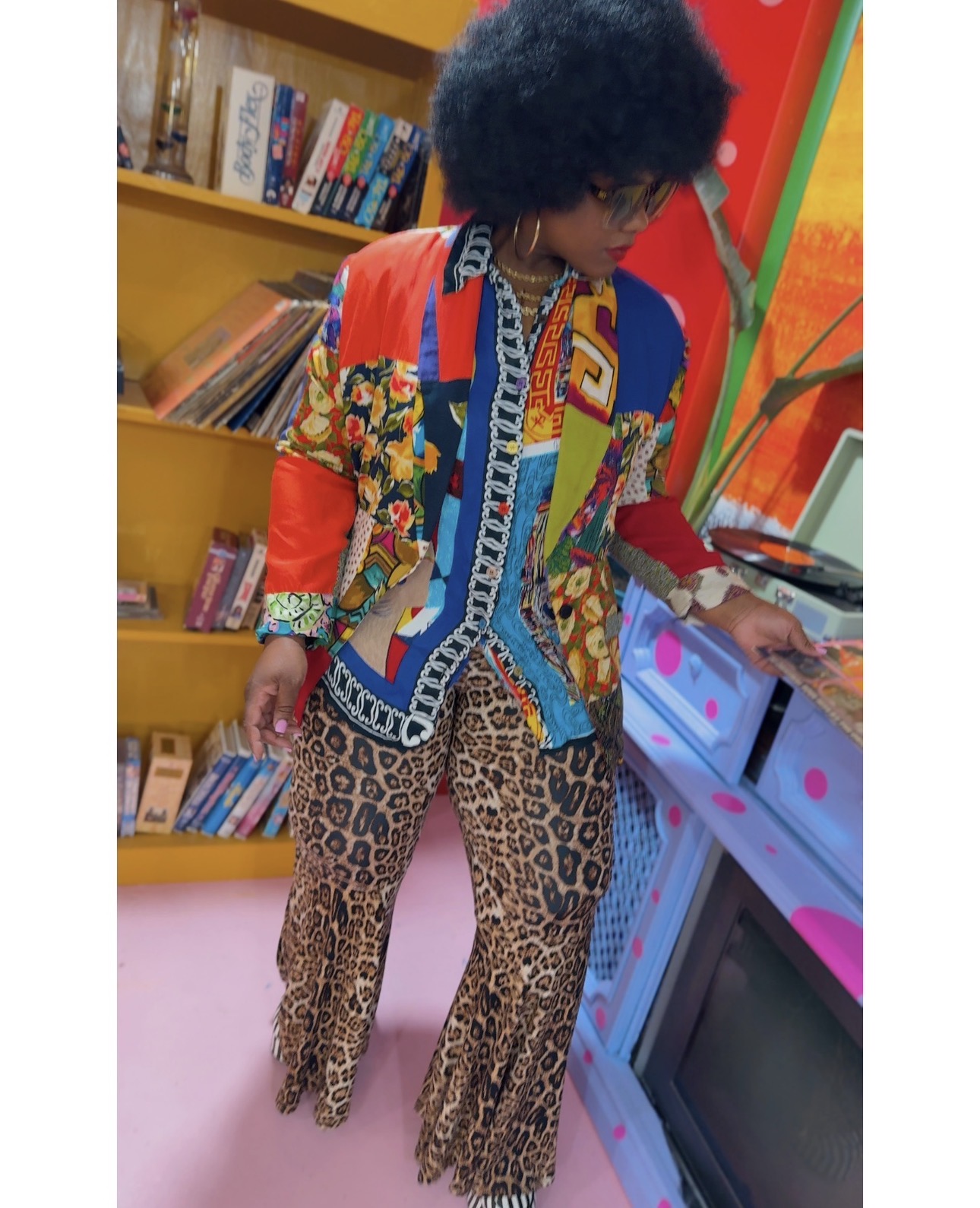 Person with curly hair wearing a colorful patterned jacket and leopard print pants, browsing books in a library.