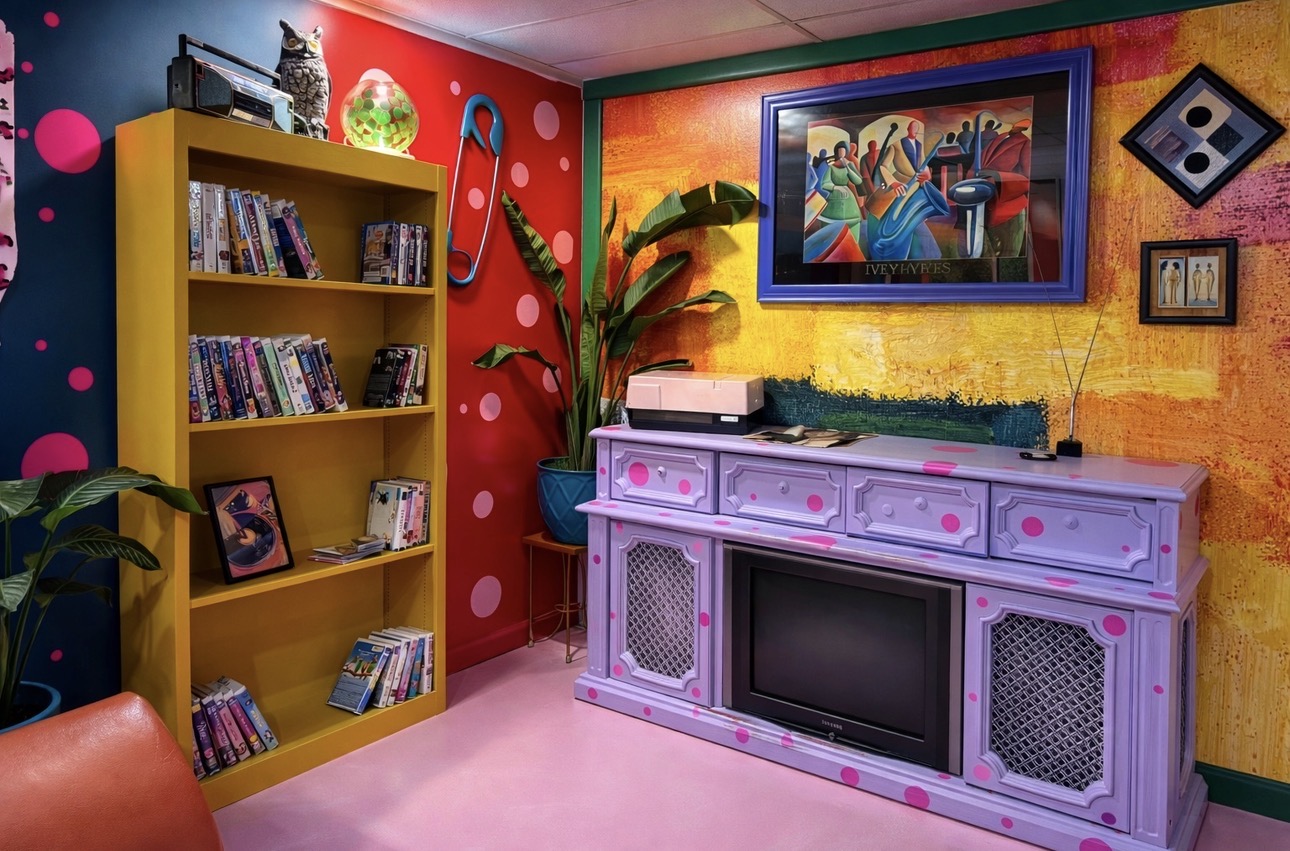 Colorful room with a yellow bookshelf, a pink and purple cabinet, a large painting, and vibrant wall colors.