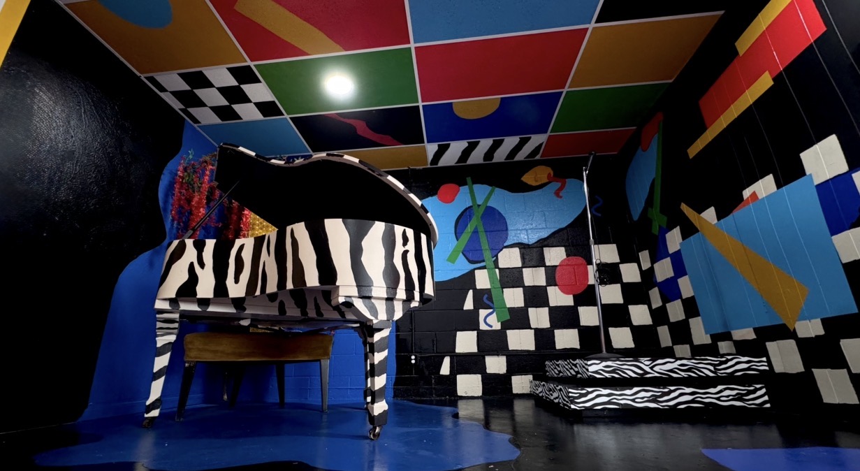 Colorful room with a zebra-striped piano, checkered walls, and abstract murals, featuring vibrant geometric patterns and bold designs.
