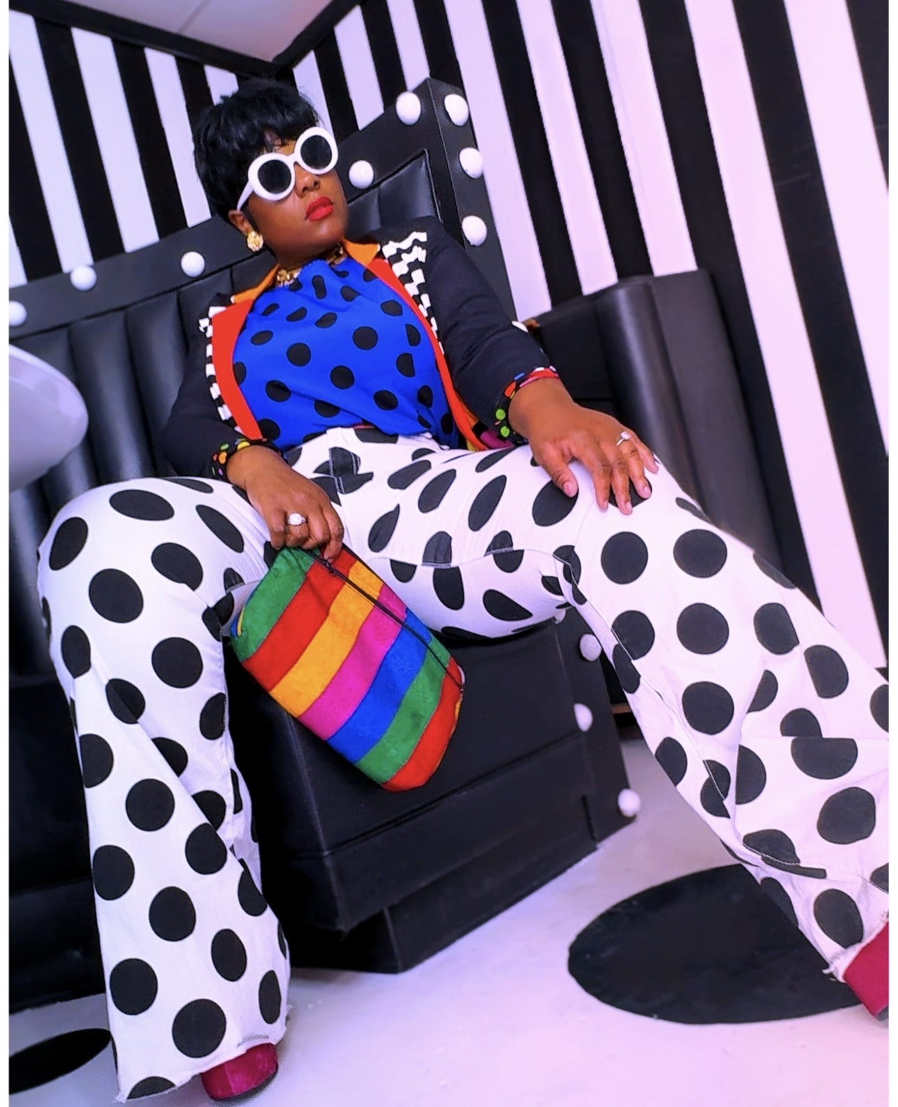 Person sitting on a black chair with black and white polka dot pants, colorful bag, and striped background.