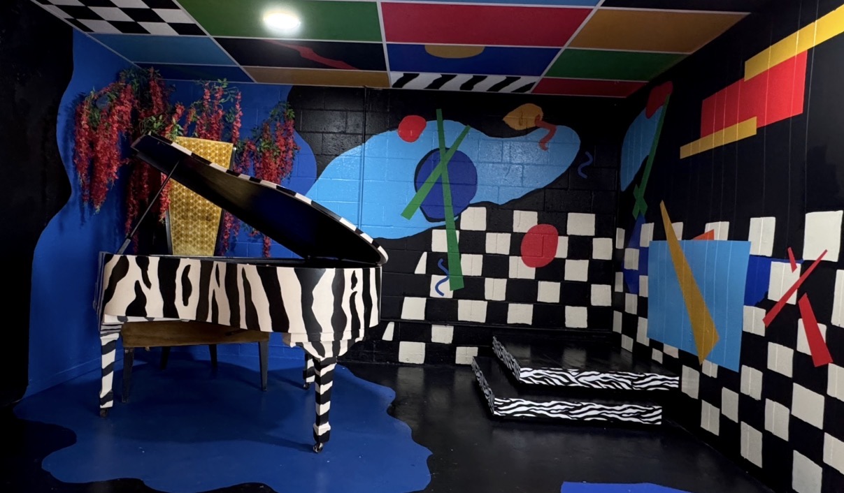 Room with colorful abstract murals, a zebra-patterned piano, and a black-and-white checkered wall, featuring vibrant geometric shapes and patterns.