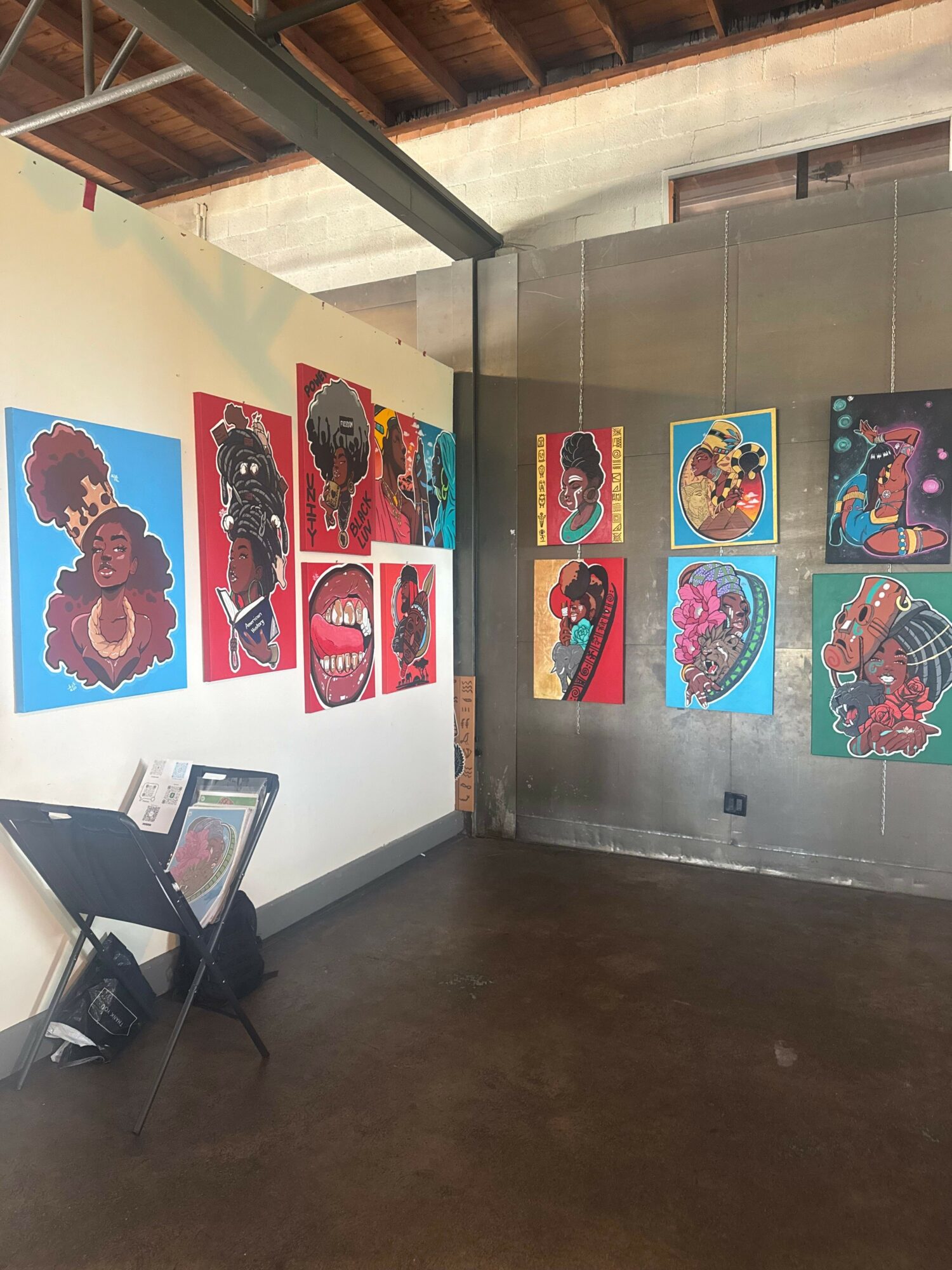 Gallery wall with colorful abstract and animal paintings, some framed, on a white wall, with a dark floor and ceiling visible.