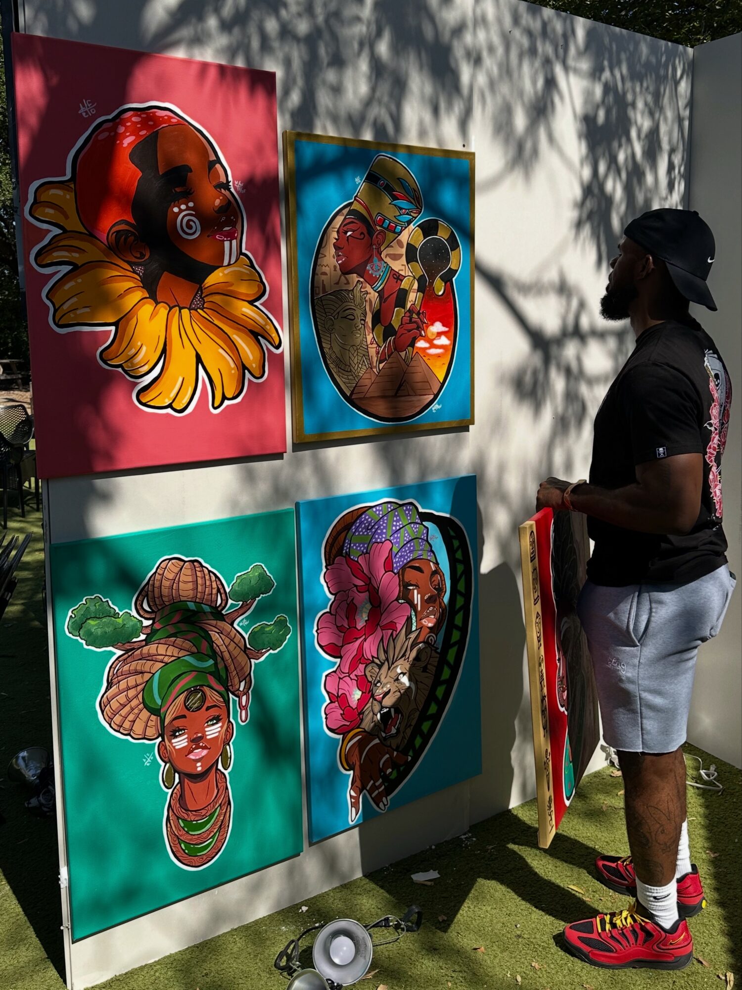 Person wearing a black cap, black shirt, white shorts, and red shoes stands outdoors near colorful artwork on a wall, with shadows cast by trees.