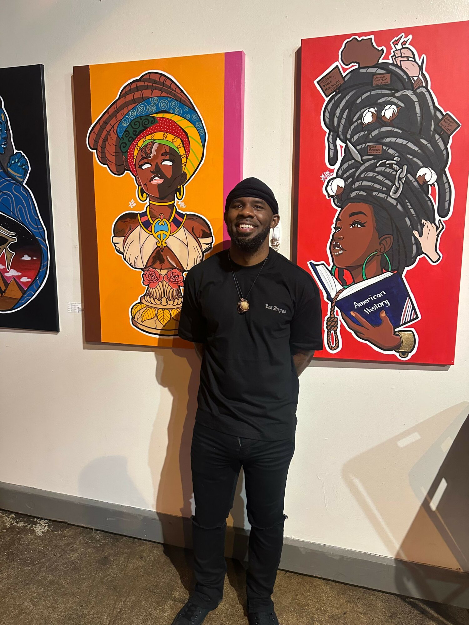 Person standing in front of colorful artwork, with one artwork featuring a woman with braided hair and glasses, and another with a woman in a hat. Alt text word count: 24. Confidence: 0.9