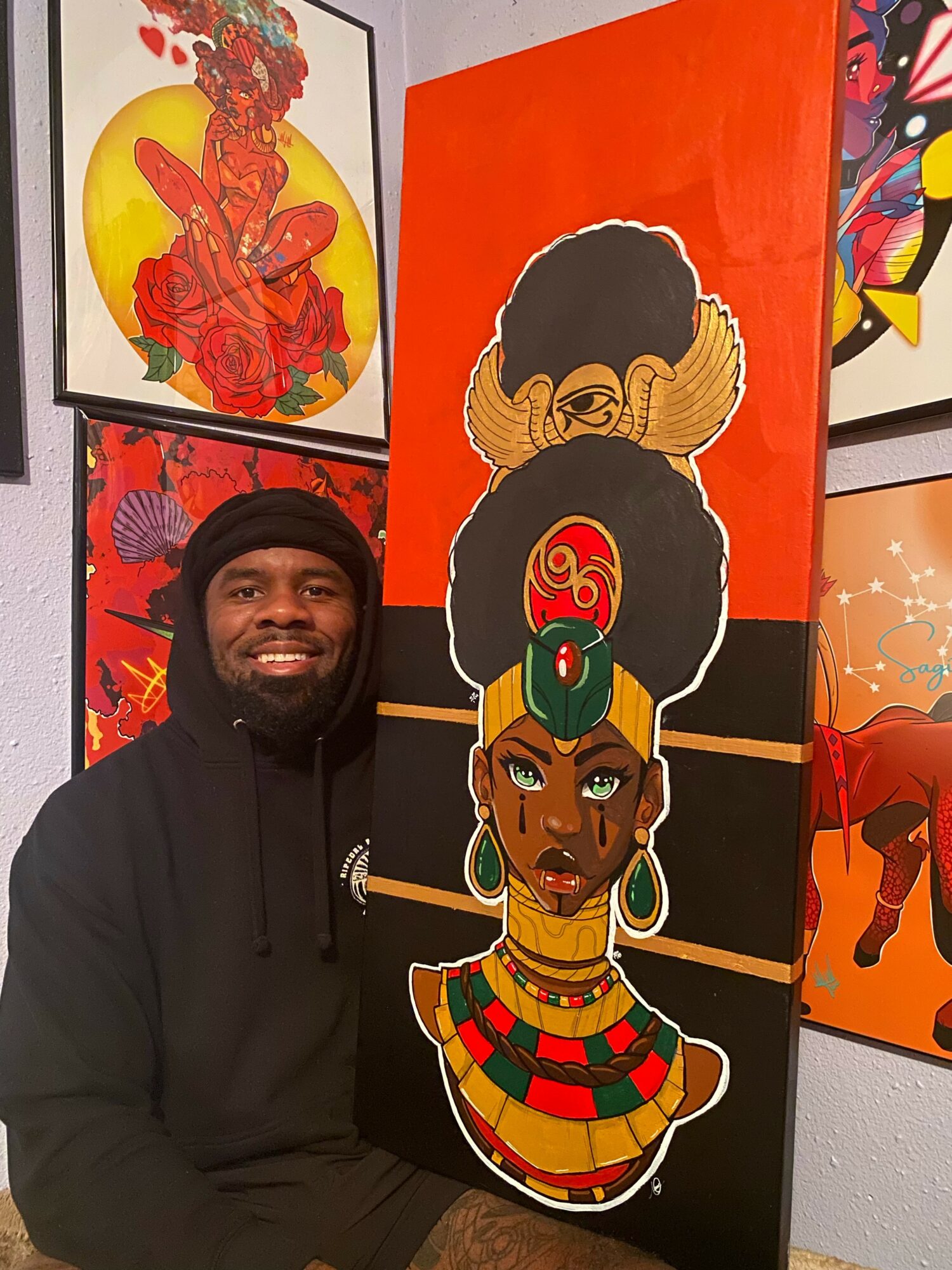 Man with a beard wearing a black hoodie, smiling, standing next to colorful African-inspired artwork featuring a woman with elaborate jewelry and headwrap.