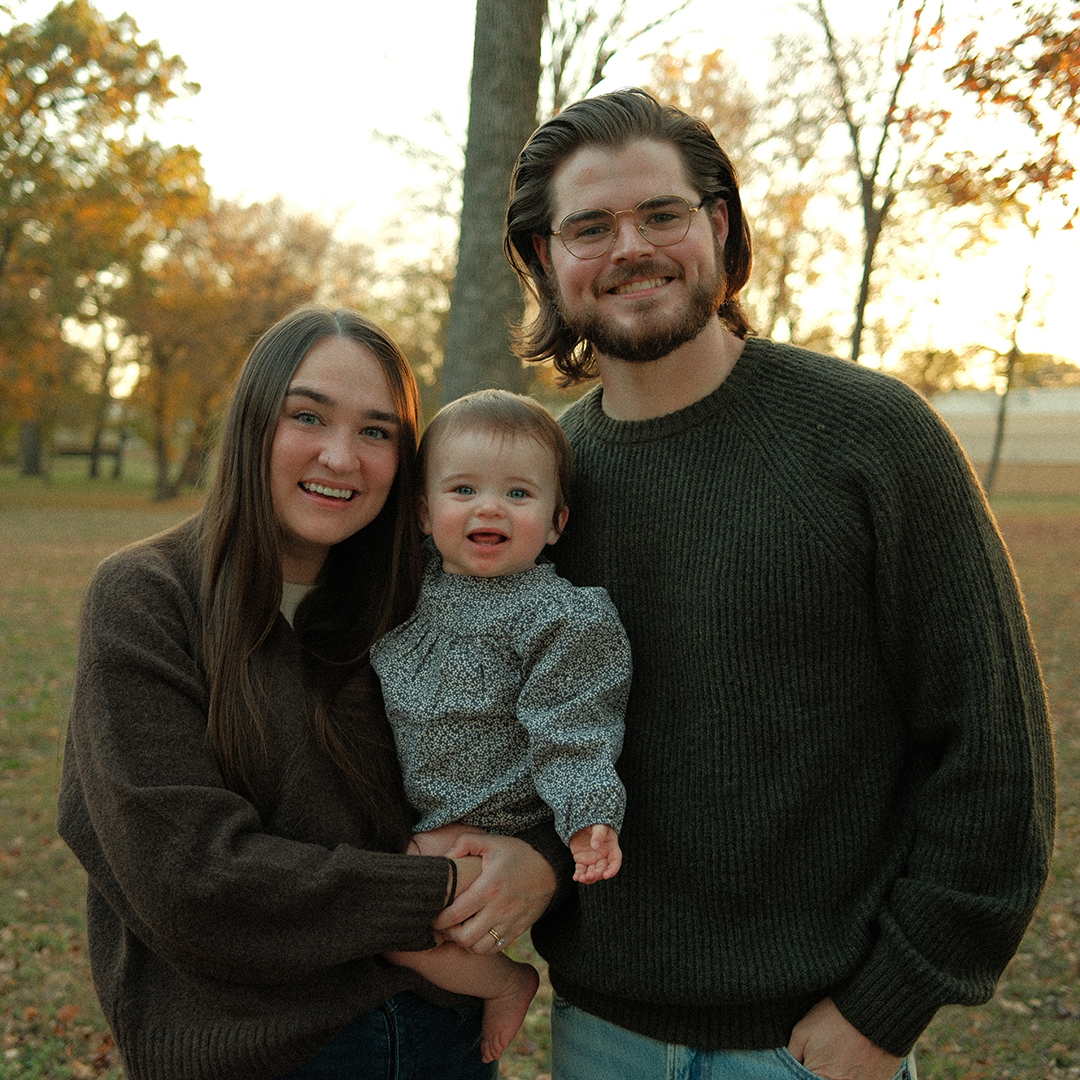 Three people outdoors in autumn, smiling, with trees and sunlight in background.