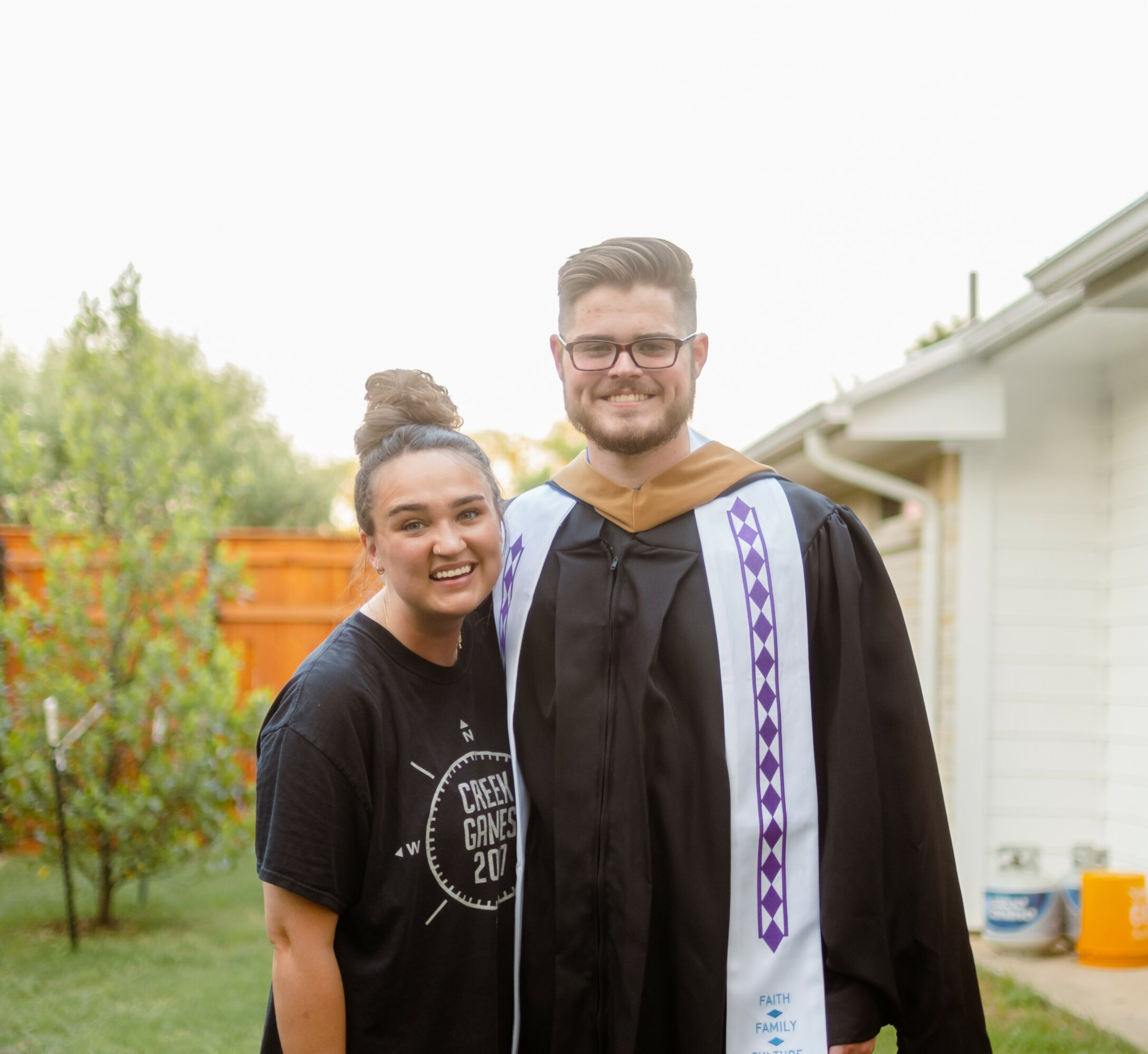 A young man in graduation gown and a young woman smiling outdoors, with trees and a house in the background.