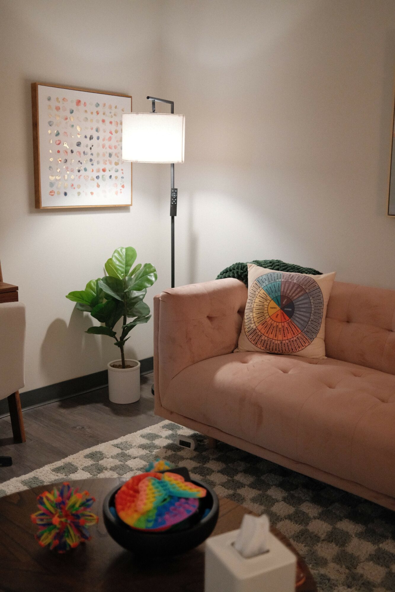 Living room with pink sofa, colorful pillow, potted plant, floor lamp, and framed artwork on white wall.