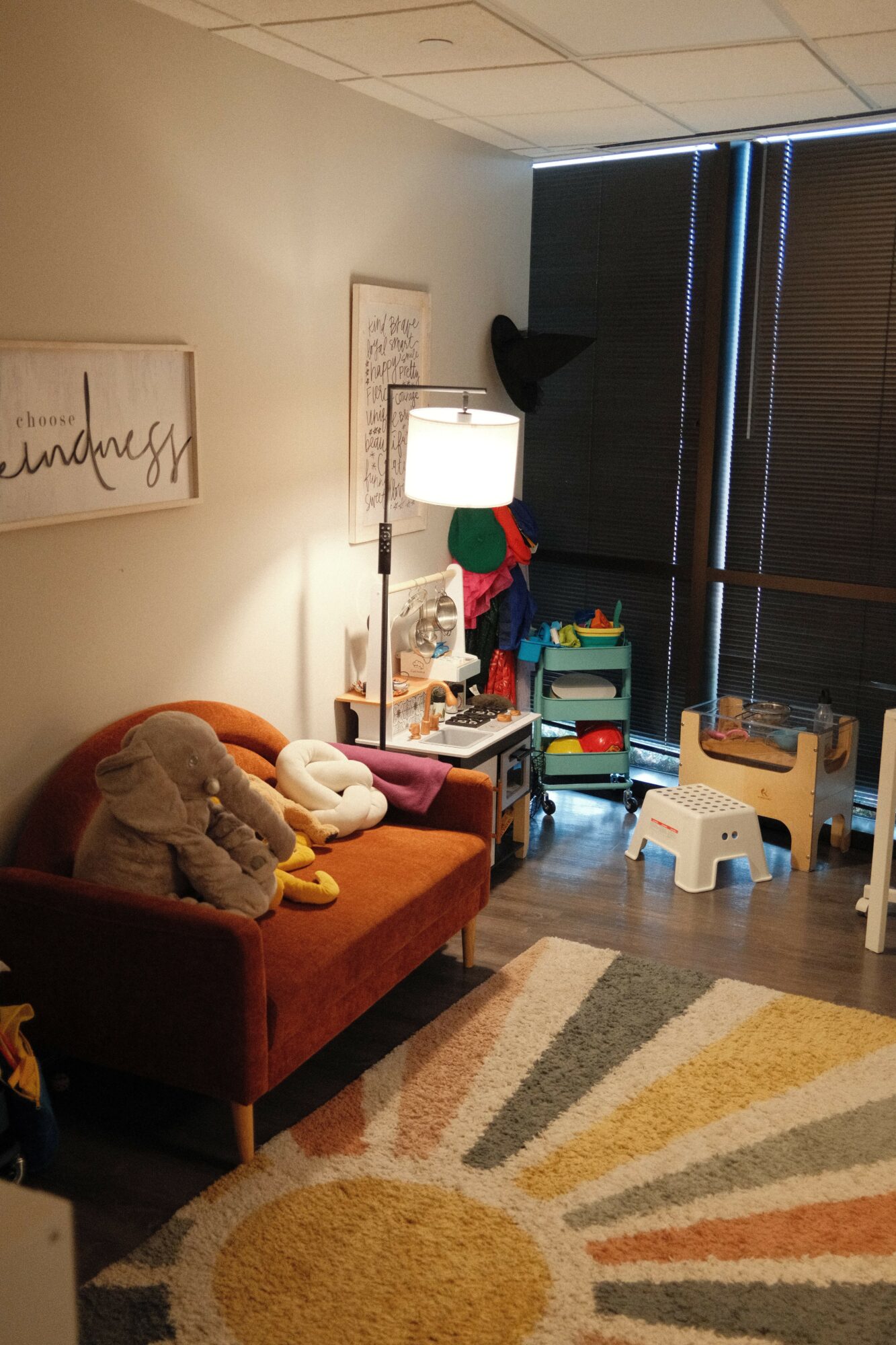 Living room with a red sofa, colorful rug, and a window with closed blinds. A lamp and various items are on a side table.