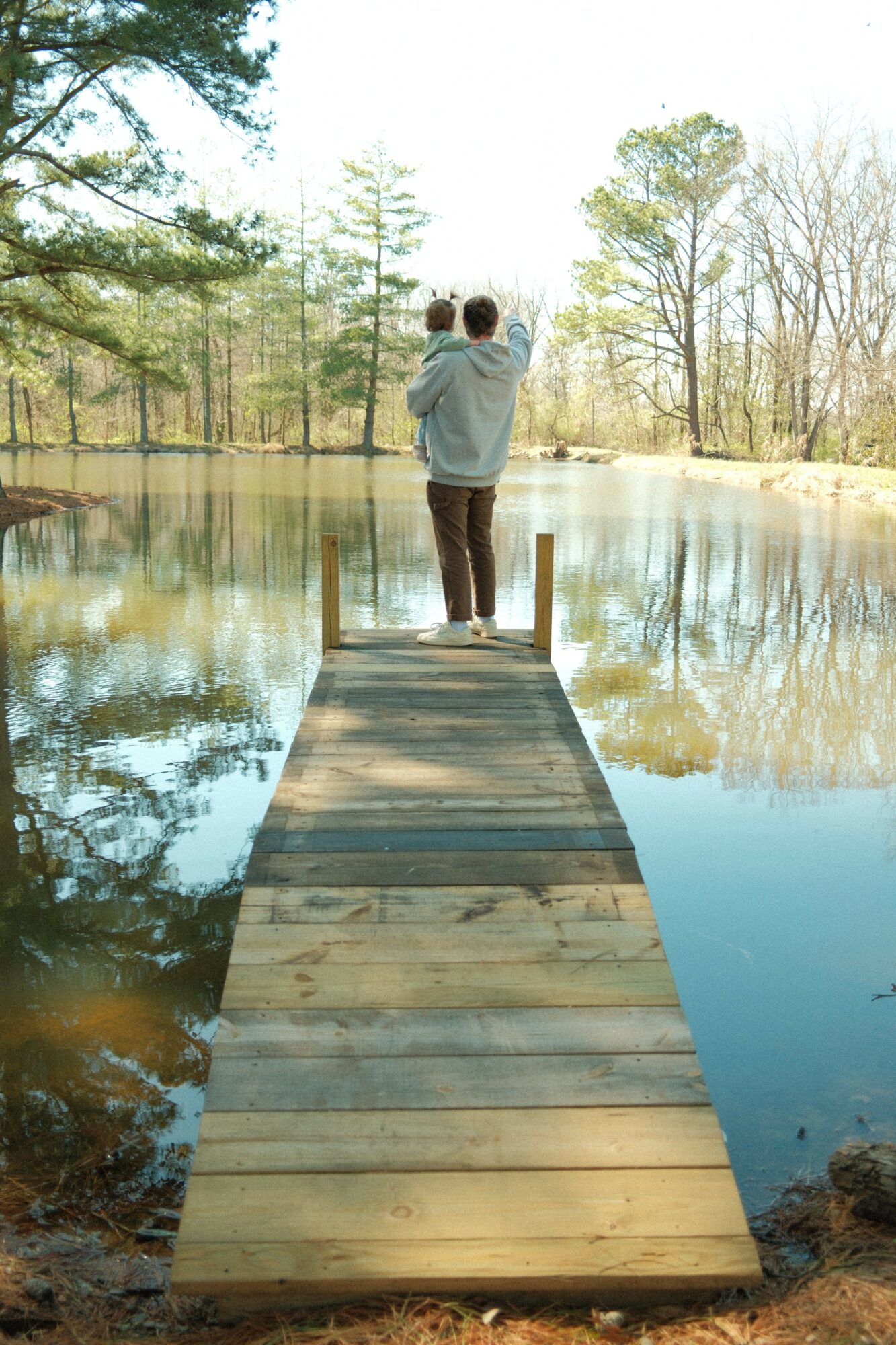 Two people walk on a wooden bridge over a calm body of water, surrounded by trees and reflections.