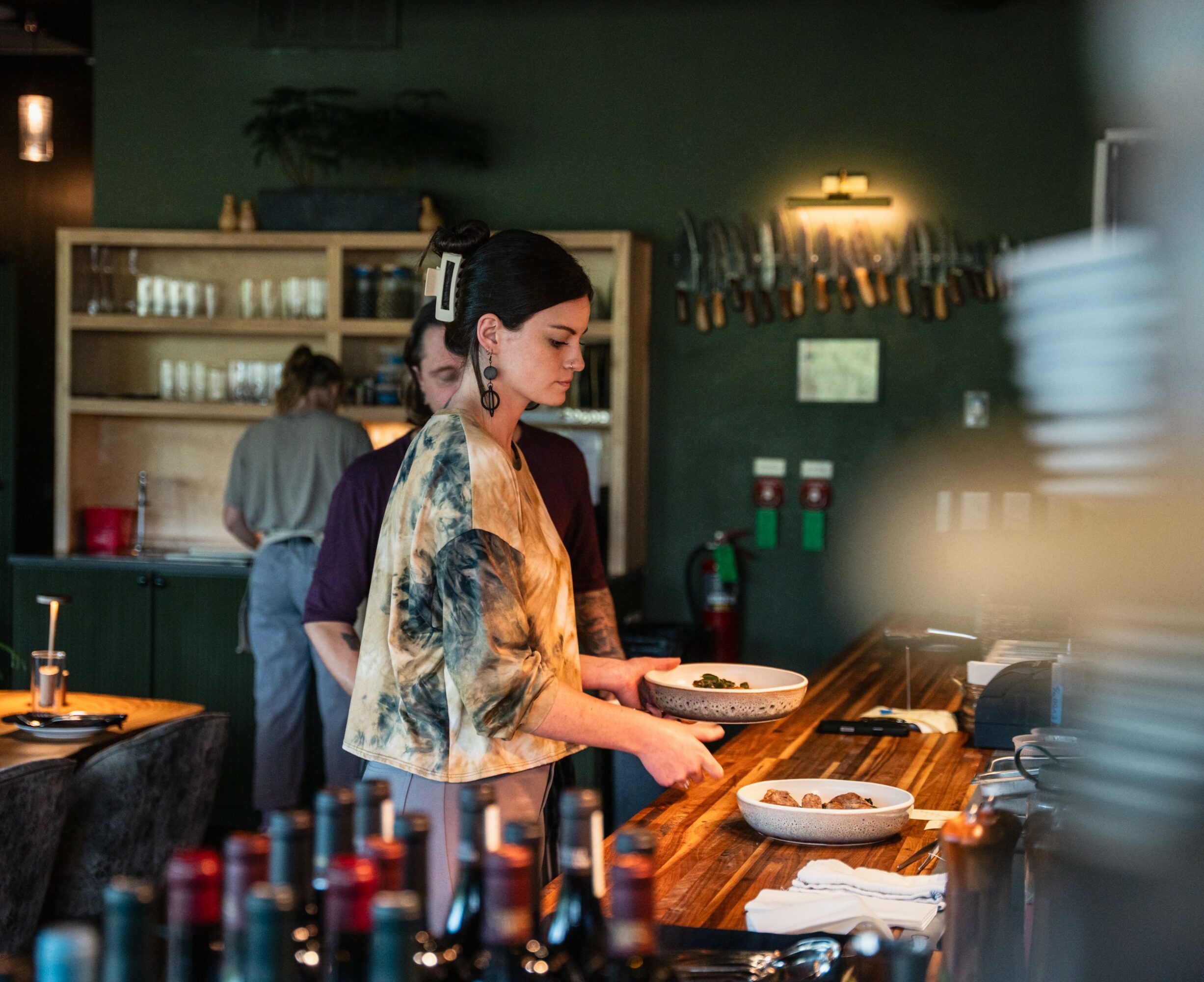 A woman in a restaurant kitchen holds a plate, with shelves, bottles, and a person in the background.