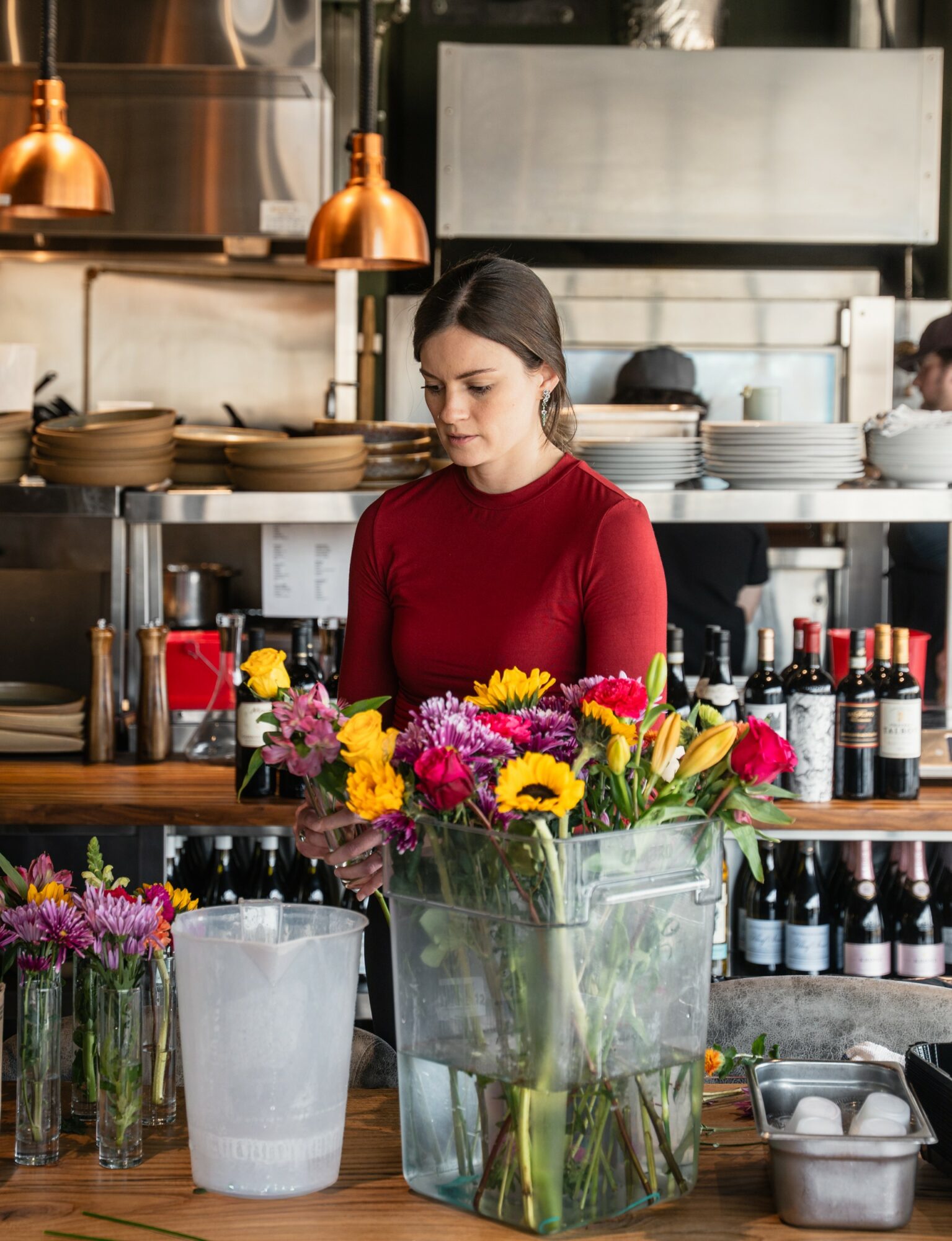 Woman in red sweater arranging a colorful flower bouquet in a glass vase in a cafe kitchen.