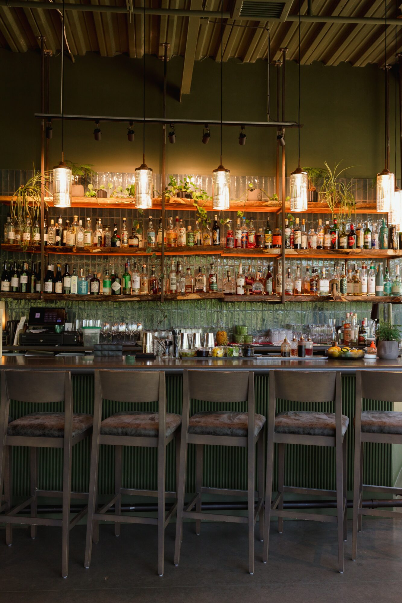 Bar counter with shelves of bottles and glasses, hanging lights, and four chairs in front, in a dimly lit setting.