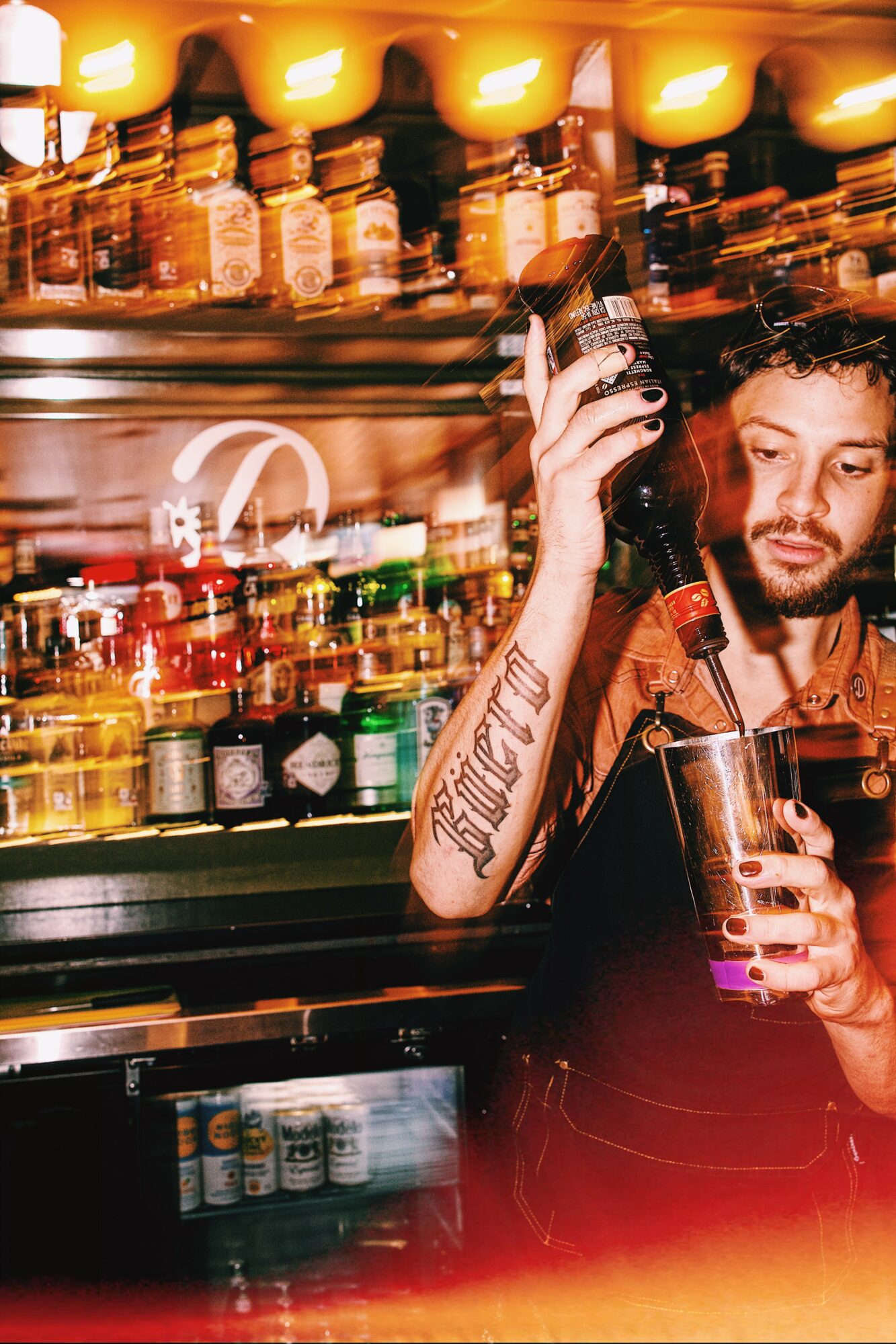Man with tattooed arm holding a bottle and glass in a bar with shelves of bottles behind him.