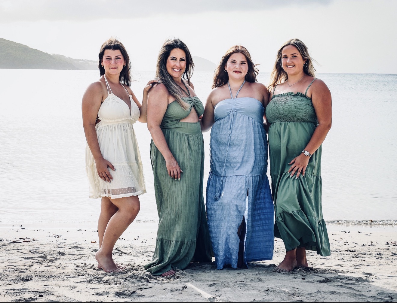Four women stand on a beach, wearing dresses, with water and sky in the background.
