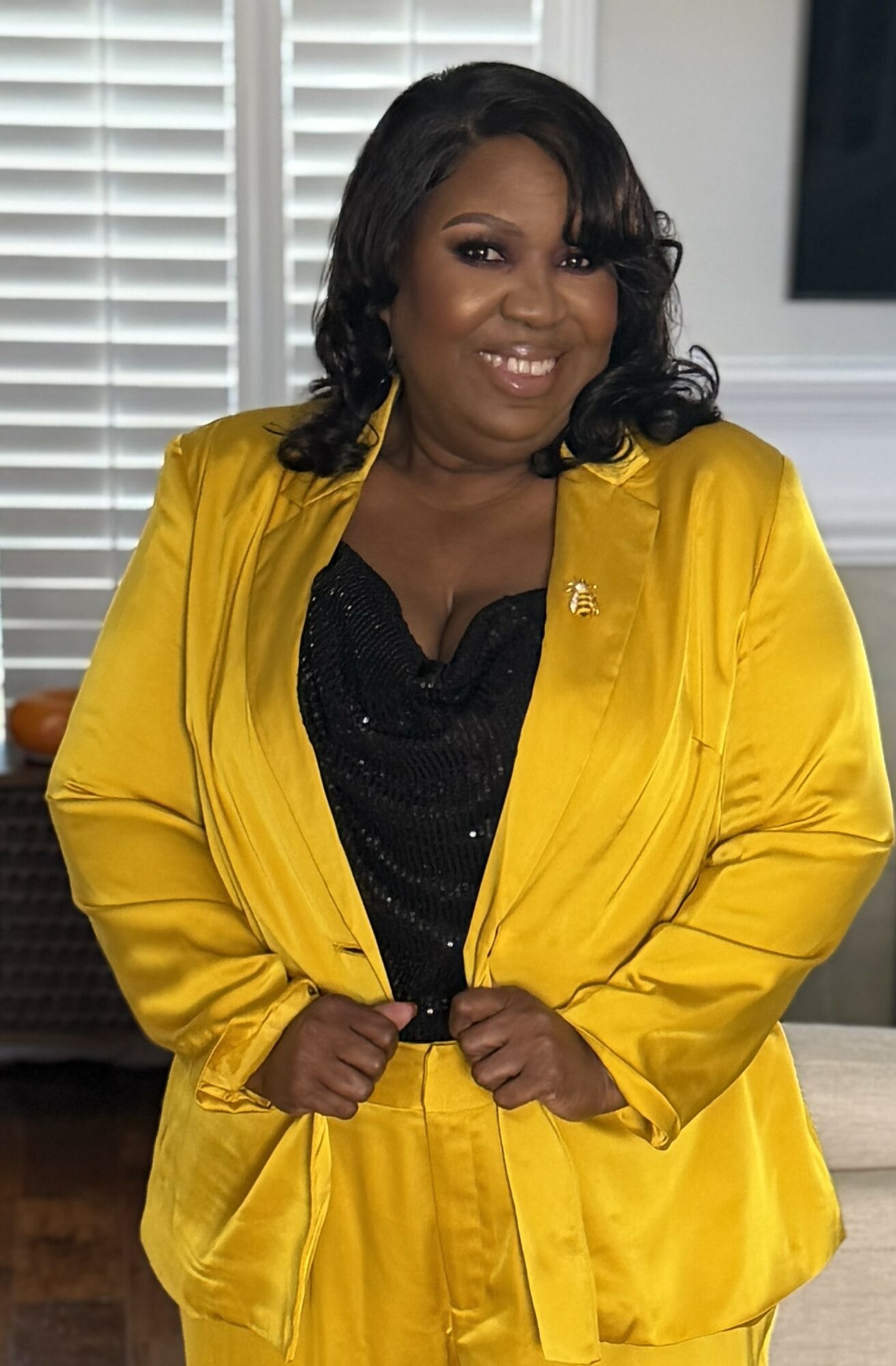Woman with black hair in a yellow blazer and black top, smiling indoors near window blinds.