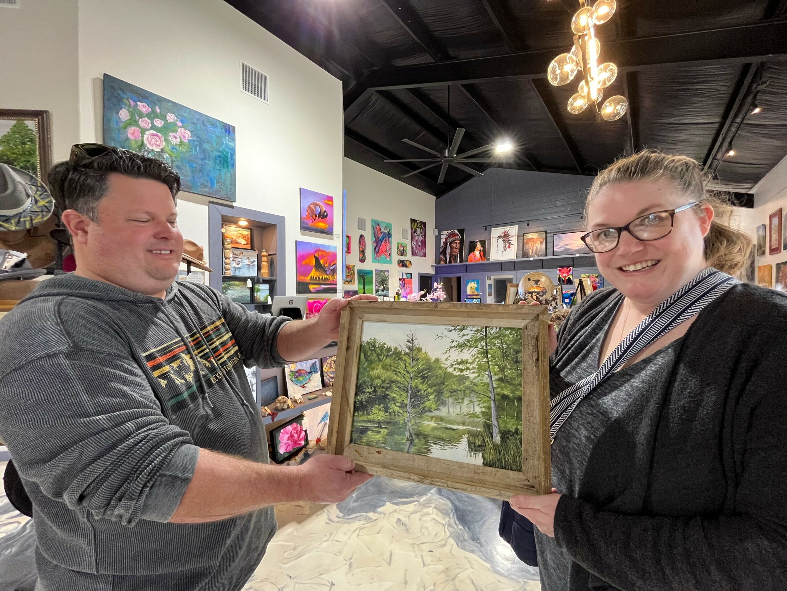 Two people holding a framed landscape painting in an art gallery, smiling, with colorful artwork on walls behind them.