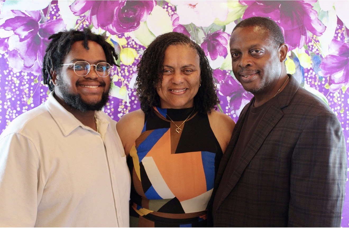 Three people standing together in front of a purple floral background, smiling, with one woman in the middle and two men on either side.