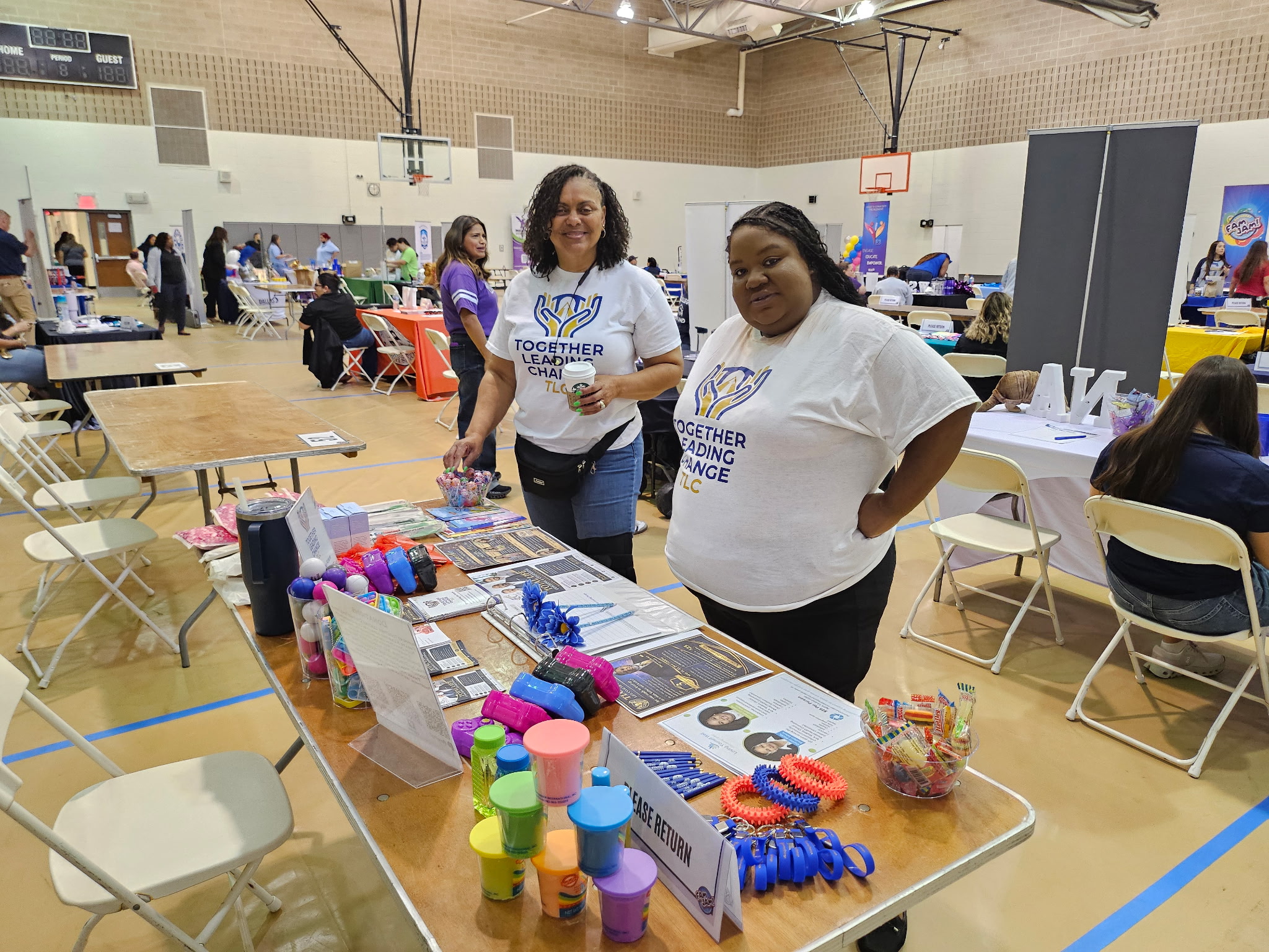 Two women standing behind a table with colorful bottles, books, and craft supplies in a large indoor event space.