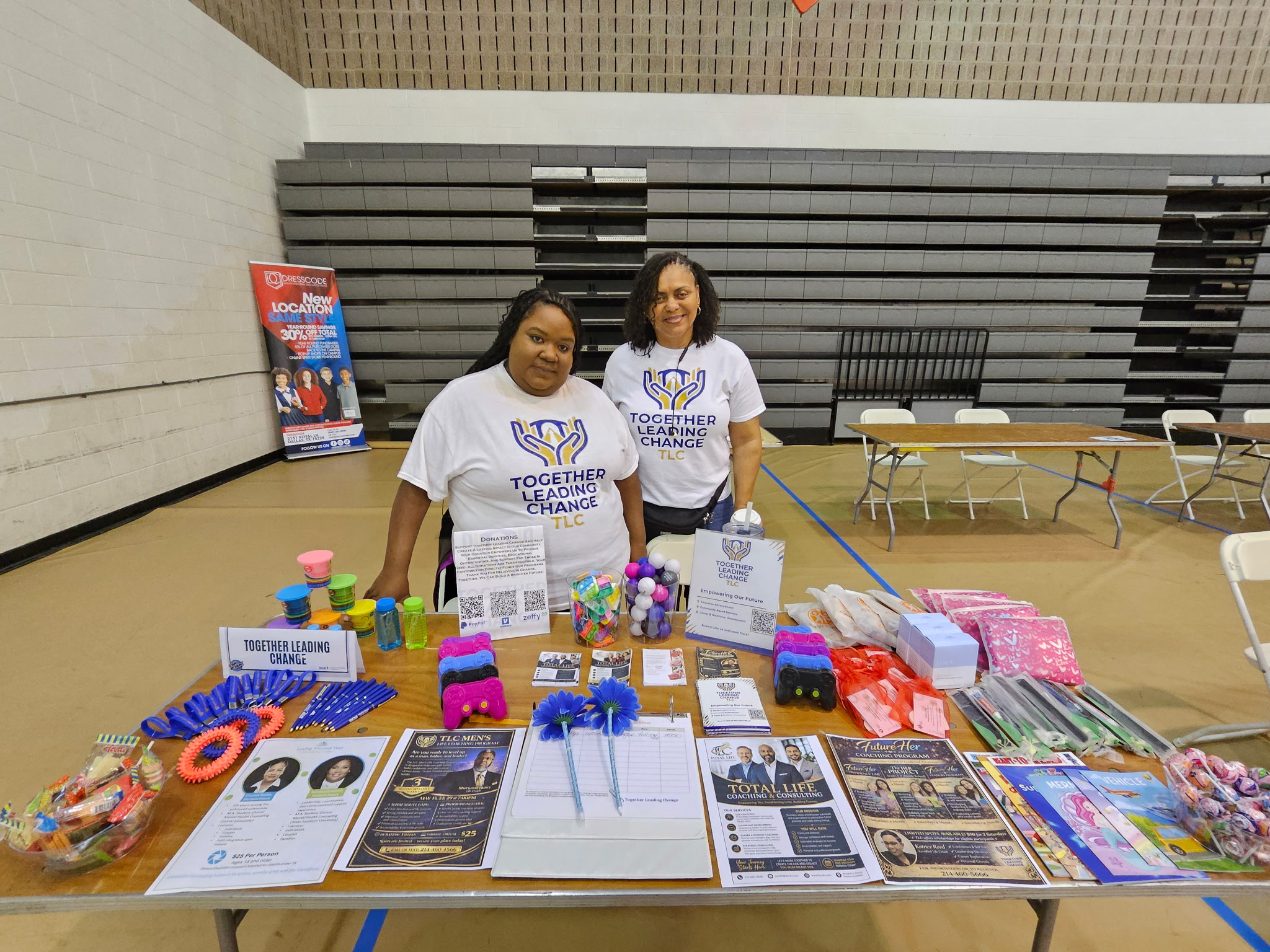 Two women stand behind a table with informational materials and colorful items in a gymnasium, smiling at the camera.