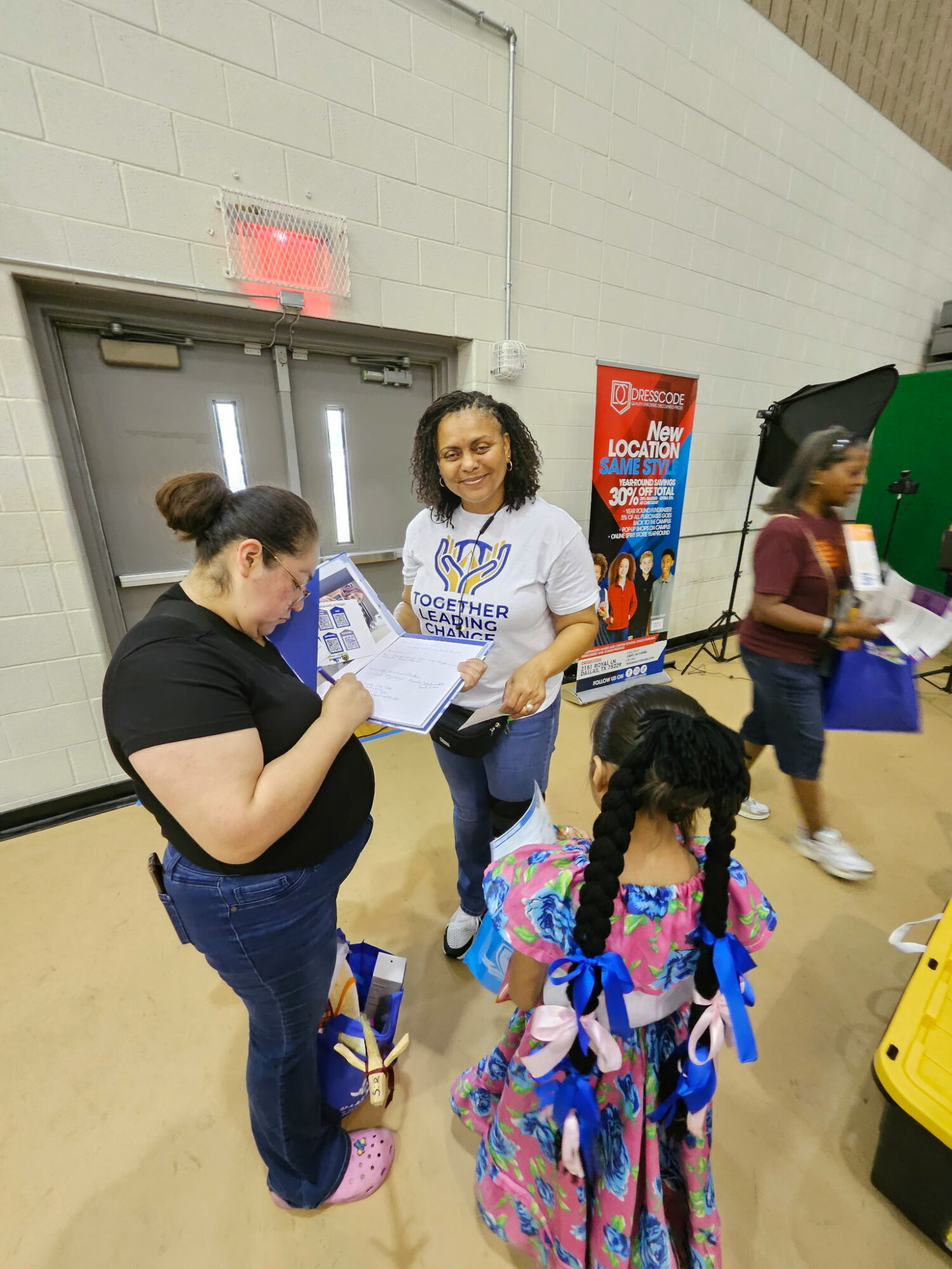 Three women and a girl in a gymnasium, one woman signing a paper, girl with braided hair and blue ribbons, background with door and posters.