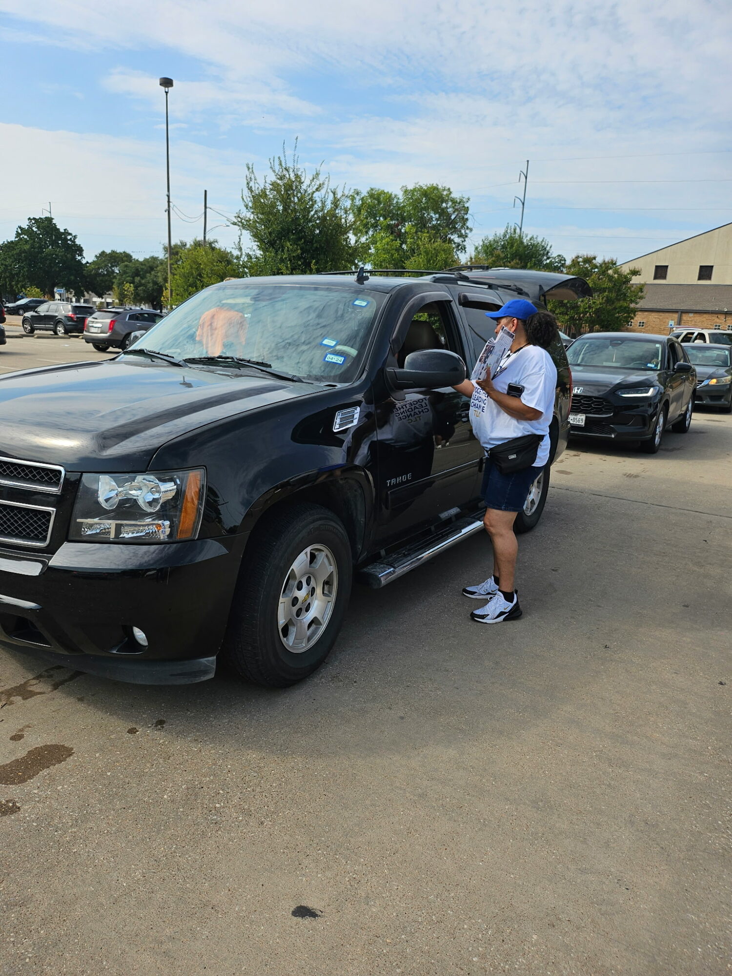 Person wearing a blue cap and white shirt interacts with a black vehicle in a parking lot, with other cars and trees in background.
