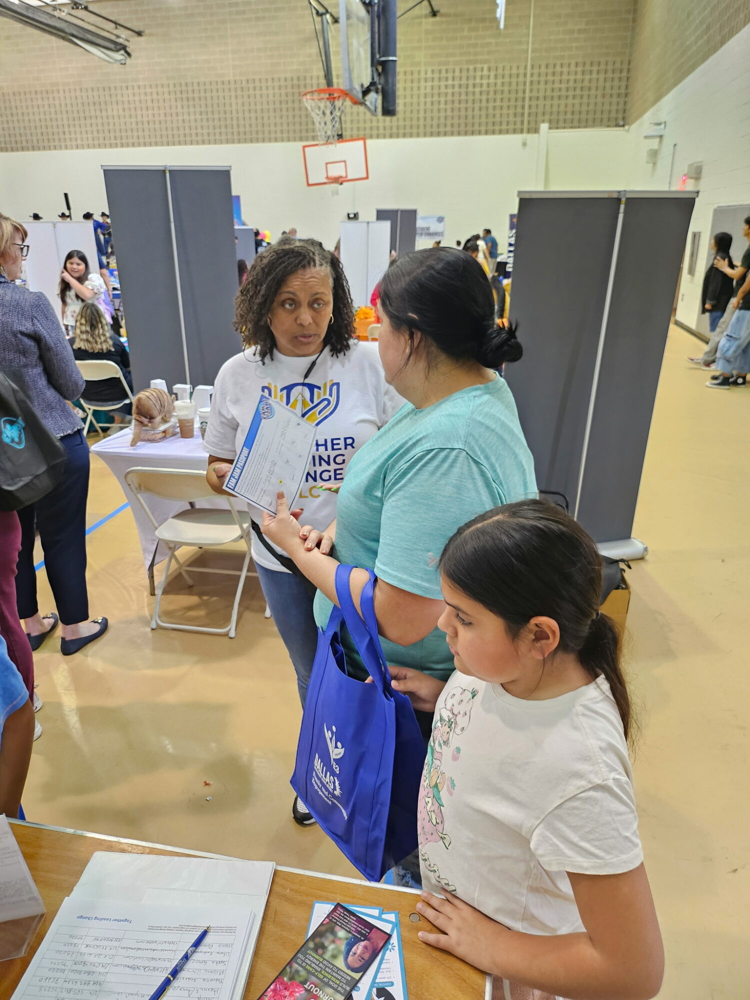 Three people, two women and a girl, engaged in conversation at an indoor event with tables and displays, some people in the background.
