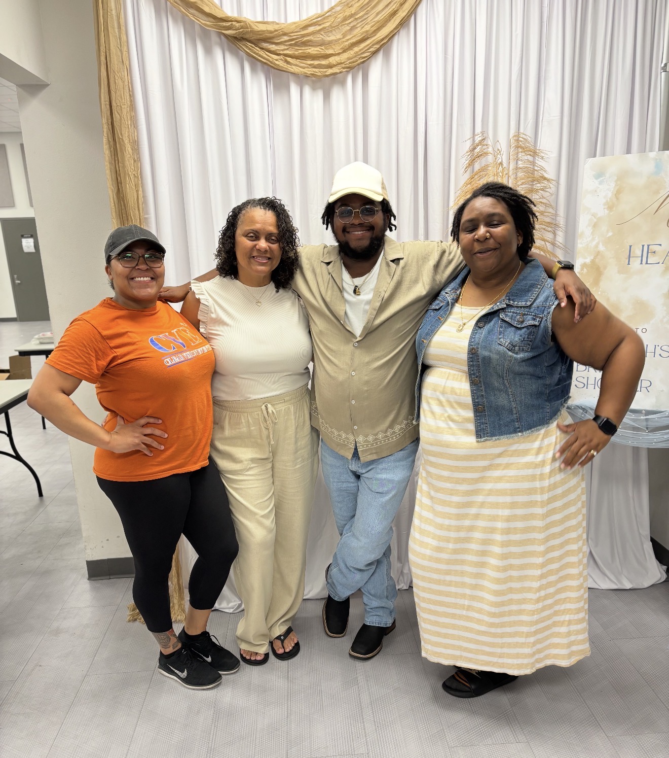 Four people standing together indoors, smiling, with a white curtain and a decorative drape behind them.