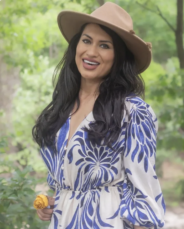 Woman smiling outdoors wearing a wide-brimmed hat and a blue and white patterned dress.