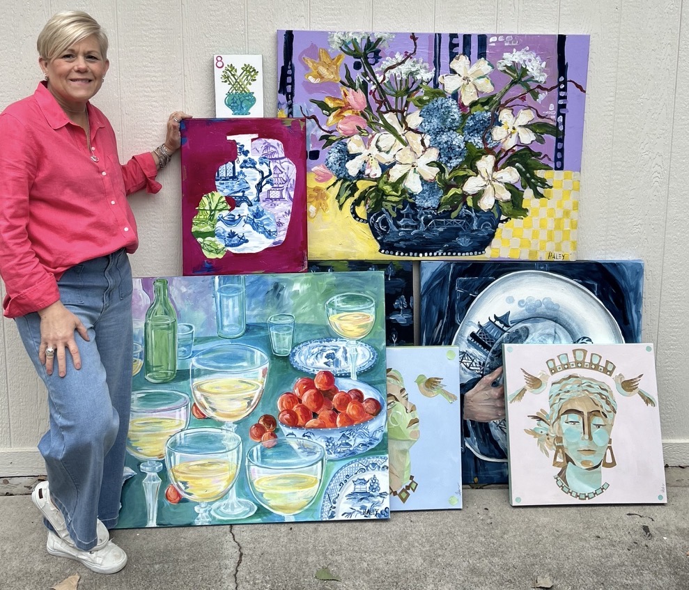 Woman in pink shirt and jeans standing next to colorful paintings of flowers, drinks, and abstract art, outdoors.