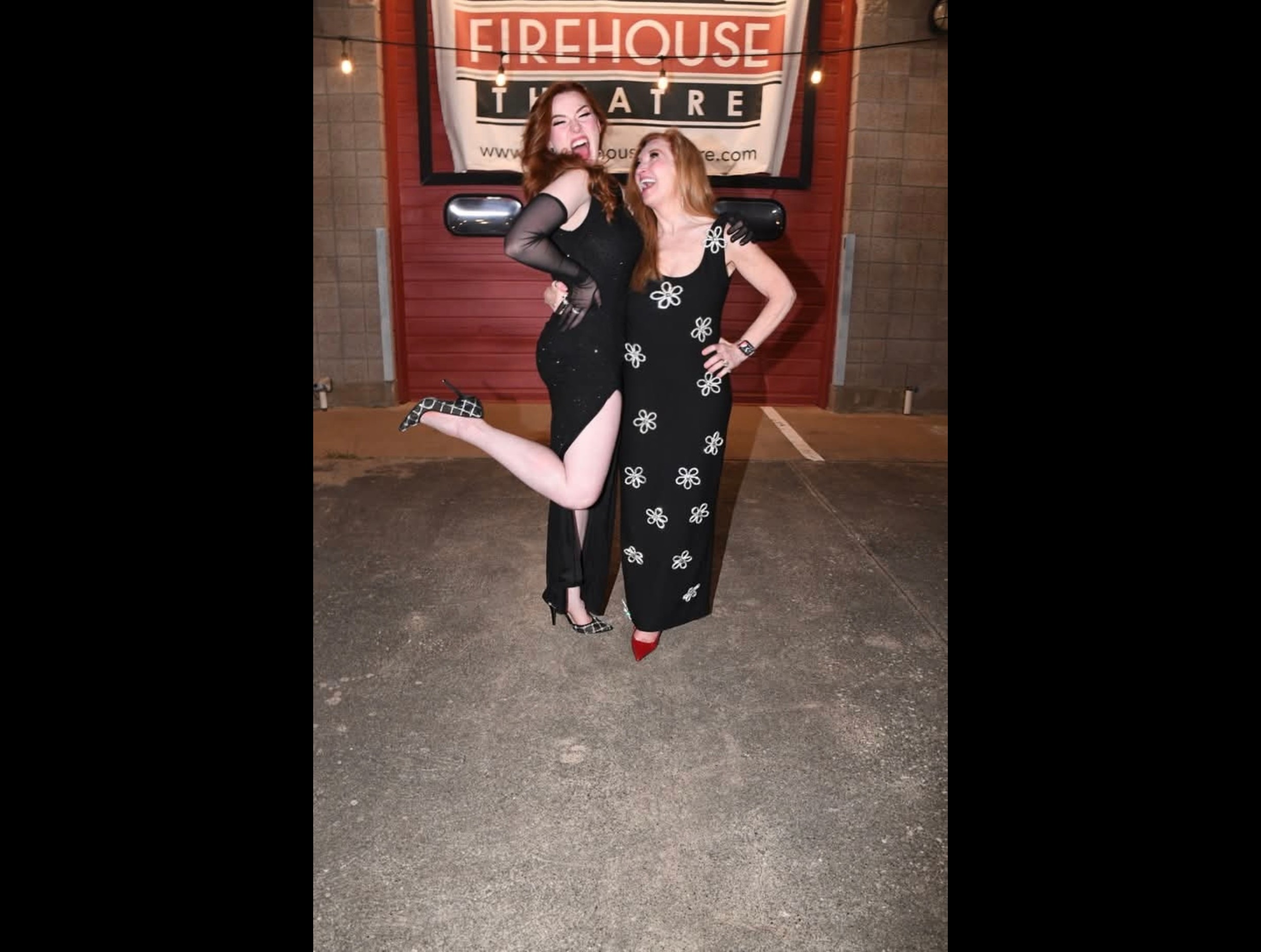 Two women pose in front of a theater entrance, one with a leg raised, both smiling, at night.