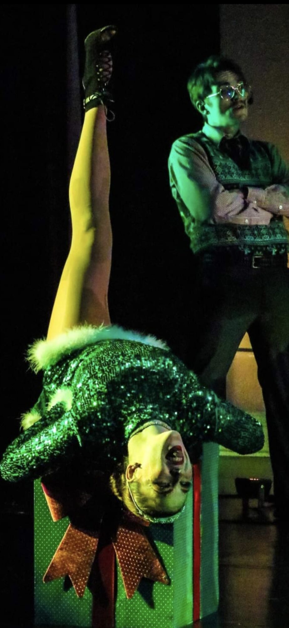 Person in a glittery costume with a bow, upside down, and another person standing with arms crossed, both on stage.